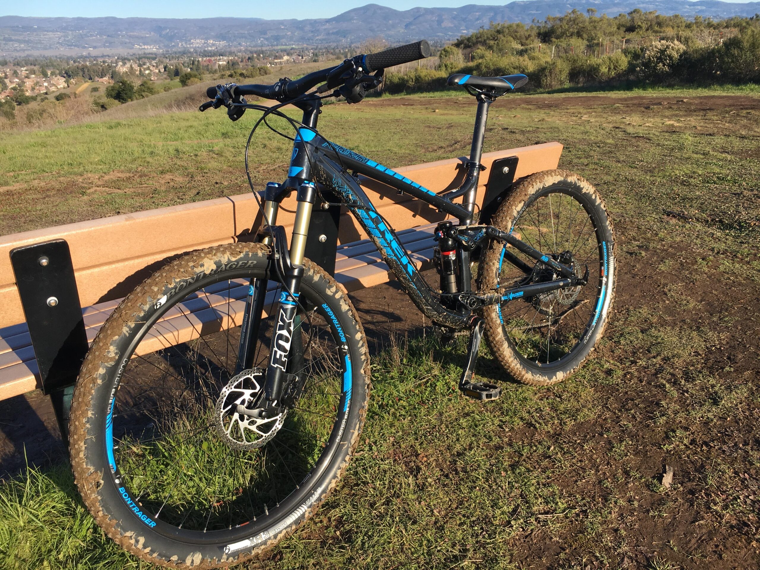 Trek Fuel EX 7: A mountain bike with black and blue accents is parked next to a wooden bench on a grassy hillside, overlooking a valley with distant mountains in the background. The bike shows signs of mud, indicating recent use on trails.