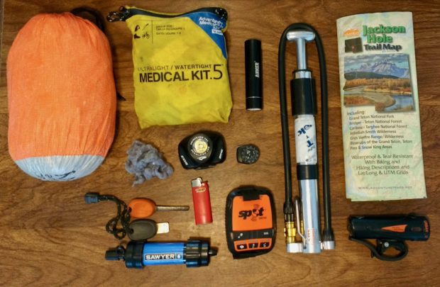 Various outdoor gear and supplies arranged on a wooden surface, including an orange compression sack, a yellow medical kit, a flashlight, a hand pump, a Jackson Hole trail map, a compass, a lighter, a set of keys, a Sawyer water filter, and a SPOT GPS device. A piece of lint or cloth is also visible among the items.