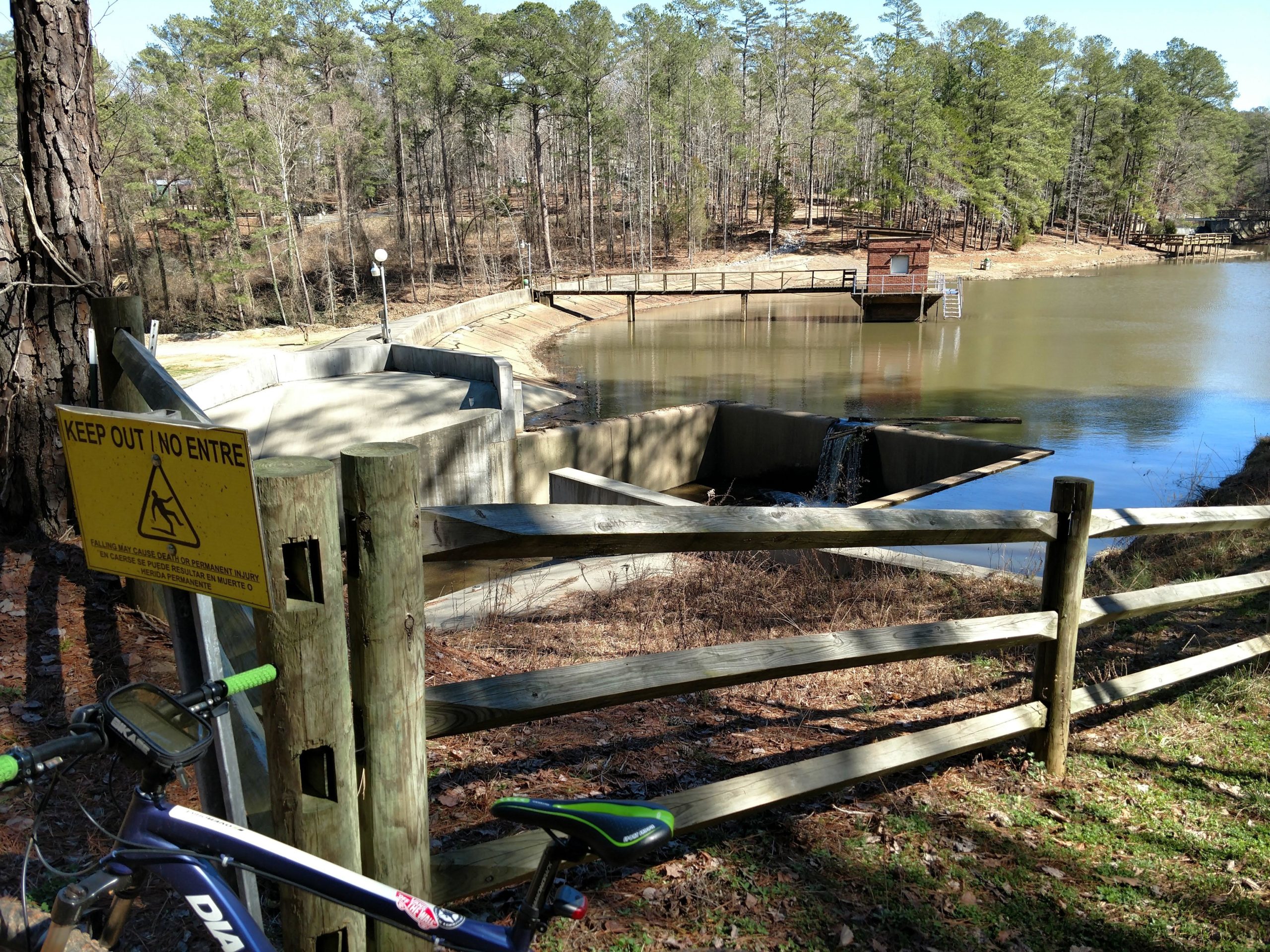 A scenic view of a calm lake surrounded by trees, featuring a concrete area with a small waterfall. A warning sign reading "KEEP OUT NO ENTREE" is visible near a wooden fence, and a bicycle is parked nearby on the grass. In the background, a wooden platform with a structure is located at the edge of the water. San-lee Park mountain bike trail.