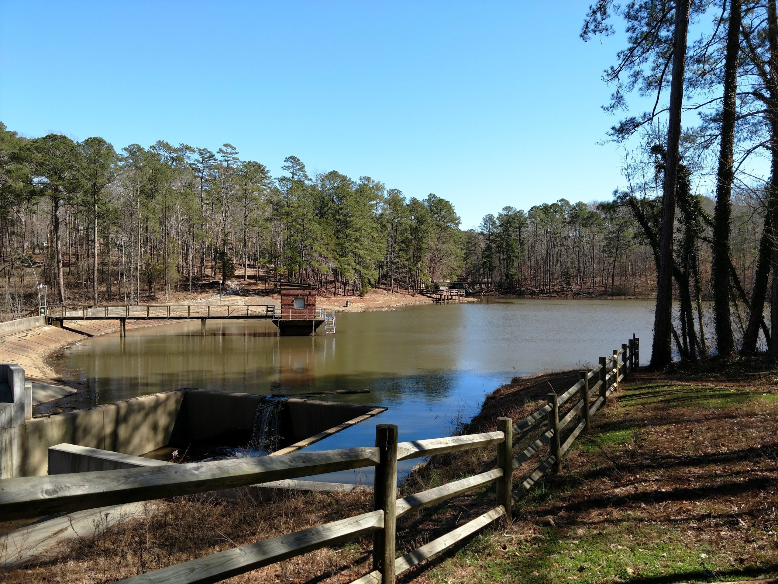 A serene lakeside scene featuring a calm water body surrounded by tall pine trees under a clear blue sky. In the foreground, a wooden fence borders the grassy area along the shore, while a wooden pier extends over the water. A small structure is visible on the pier, and a spillway can be seen at the water's edge. The landscape is peaceful, suggesting a natural setting for relaxation or recreation. San-lee Park mountain bike trail.