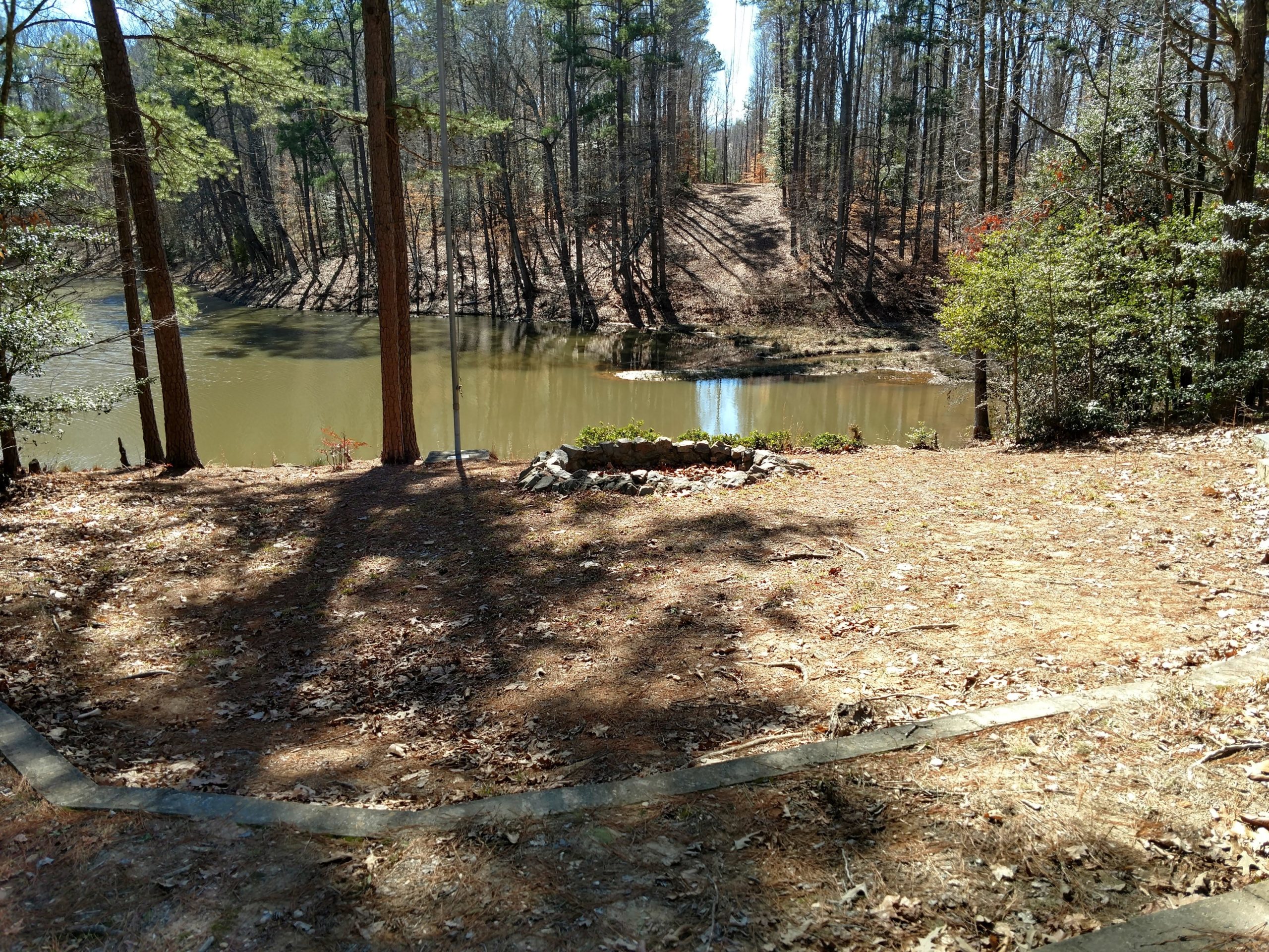 A serene view of a tranquil pond surrounded by trees, with a grassy area in the foreground. Sunlight filters through the branches, casting shadows on the ground, while the water reflects the natural scenery. The background features a gently sloping path leading away from the pond. San-lee Park mountain bike trail.