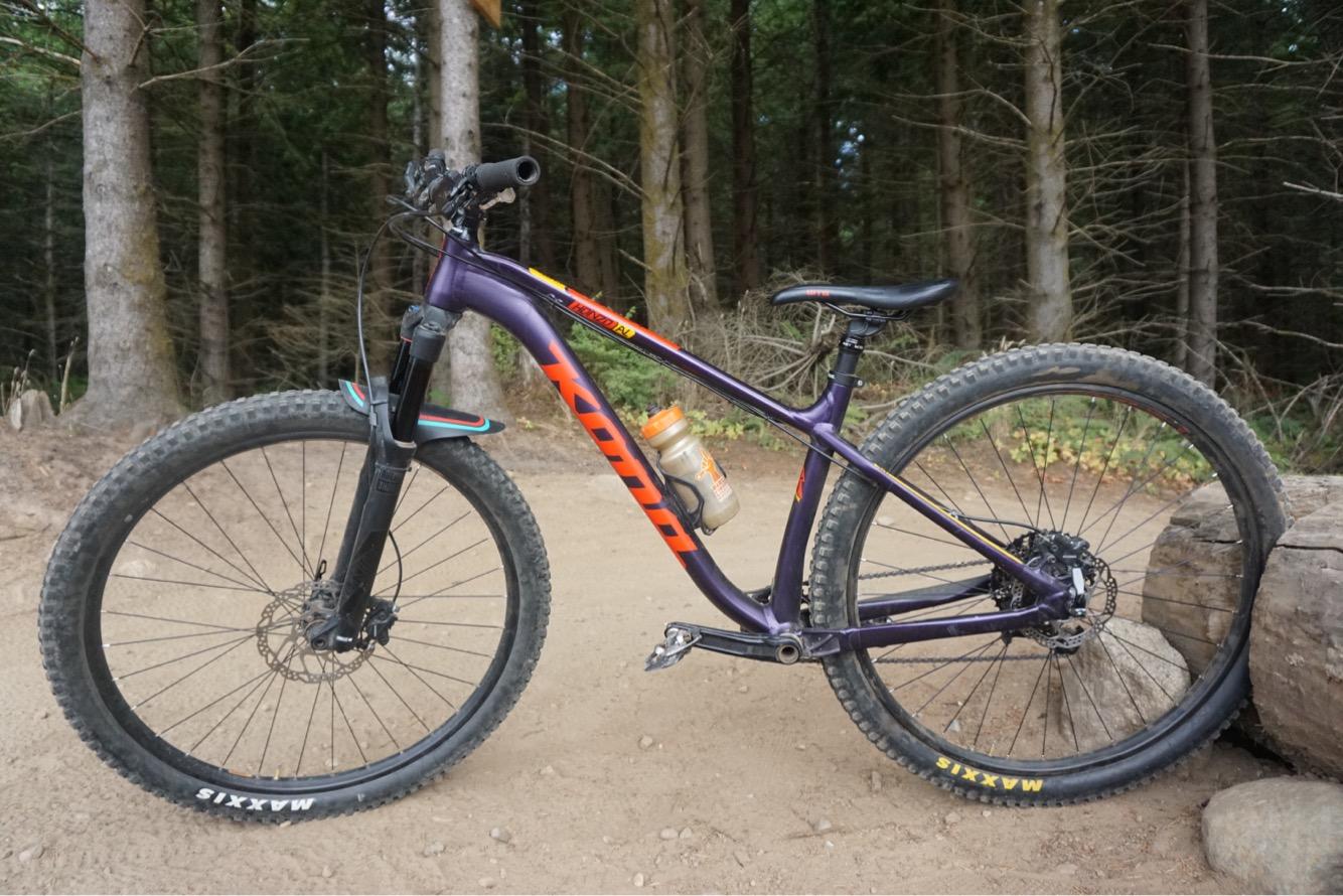 Kona Honzo: A purple mountain bike with a sturdy frame and knobby tires, positioned on a sandy trail surrounded by trees. The bike has a water bottle attached and features a suspension fork. The background shows a natural wooded area.