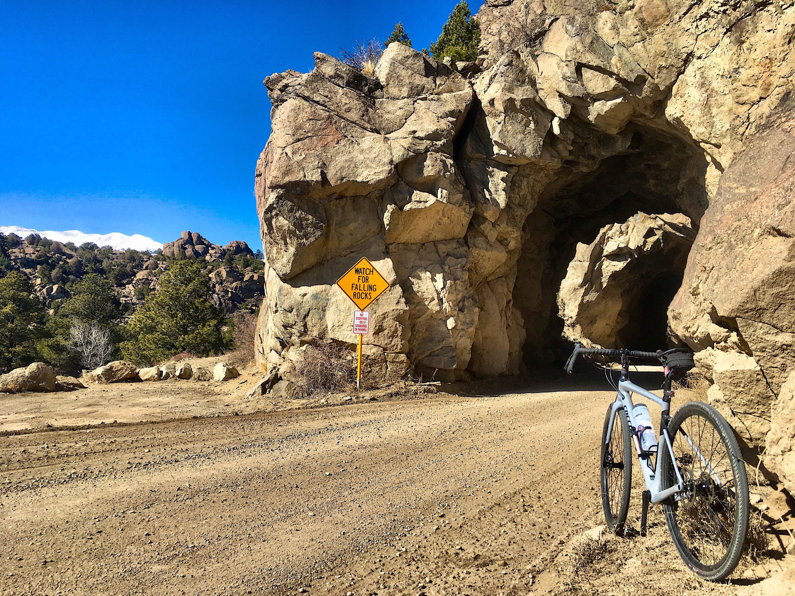 A mountain bike stands next to a rocky tunnel on a dirt road, with a yellow warning sign that reads "WATCH FOR FALLING ROCKS." The sky is clear blue, and rocky hills can be seen in the background, with snow-capped peaks visible on the horizon. River Road / #371 mountain bike trail.