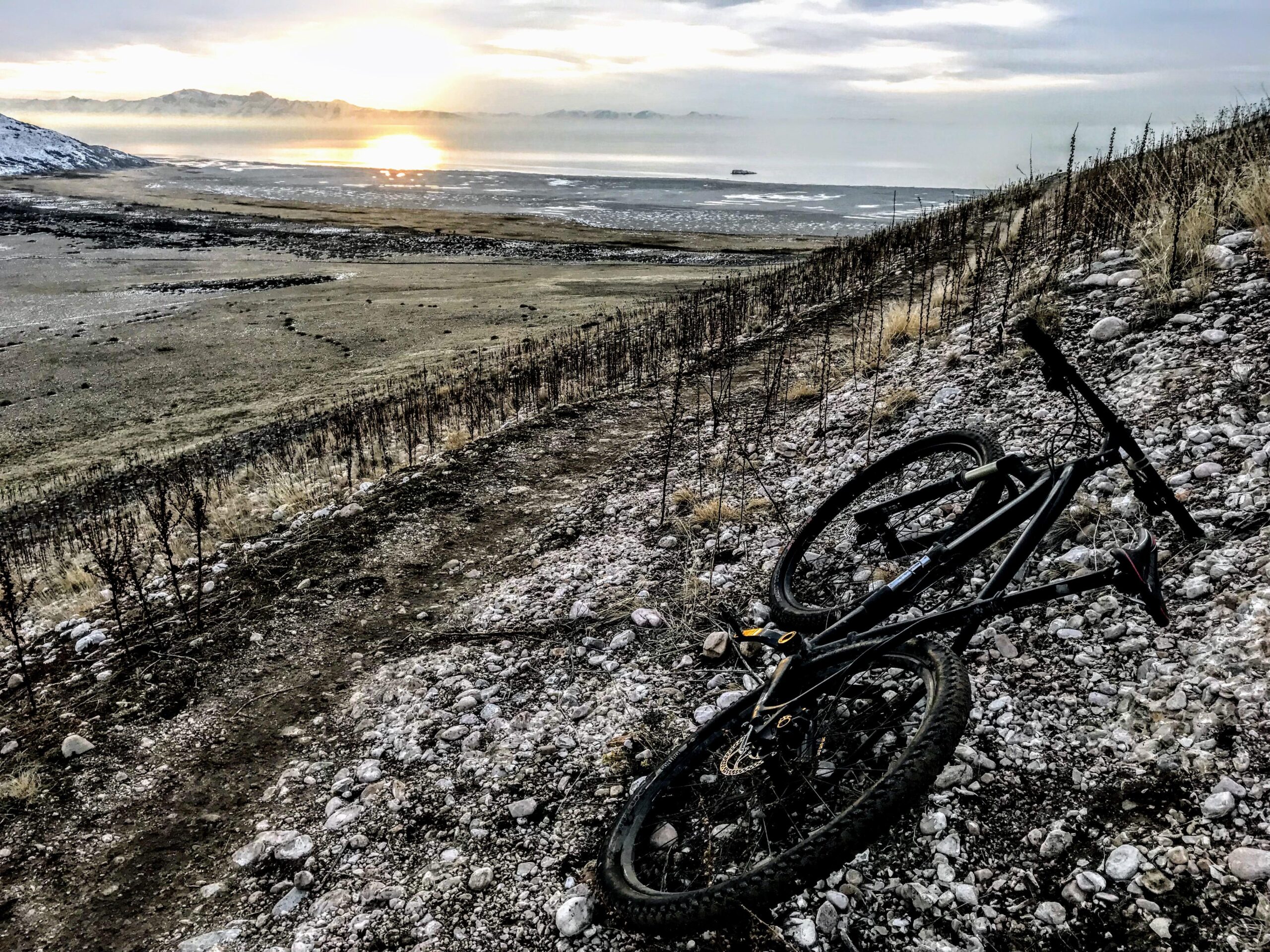 Specialized Stumpjumper Comp 29er: A mountain bike rests on a rocky hillside overlooking a vast landscape at sunset, with the sun reflecting off a distant body of water and mountains visible in the background. The scene captures a serene and adventurous outdoor atmosphere.