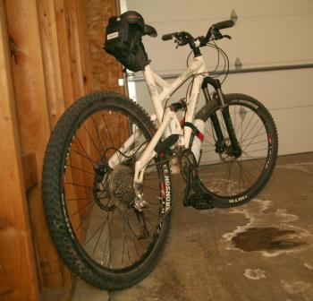 Airborne Zeppelin Elite: A white mountain bike with thick tires, positioned against a wooden wall in a garage, with a small black bag mounted on the handlebars. The garage door is partially visible in the background.