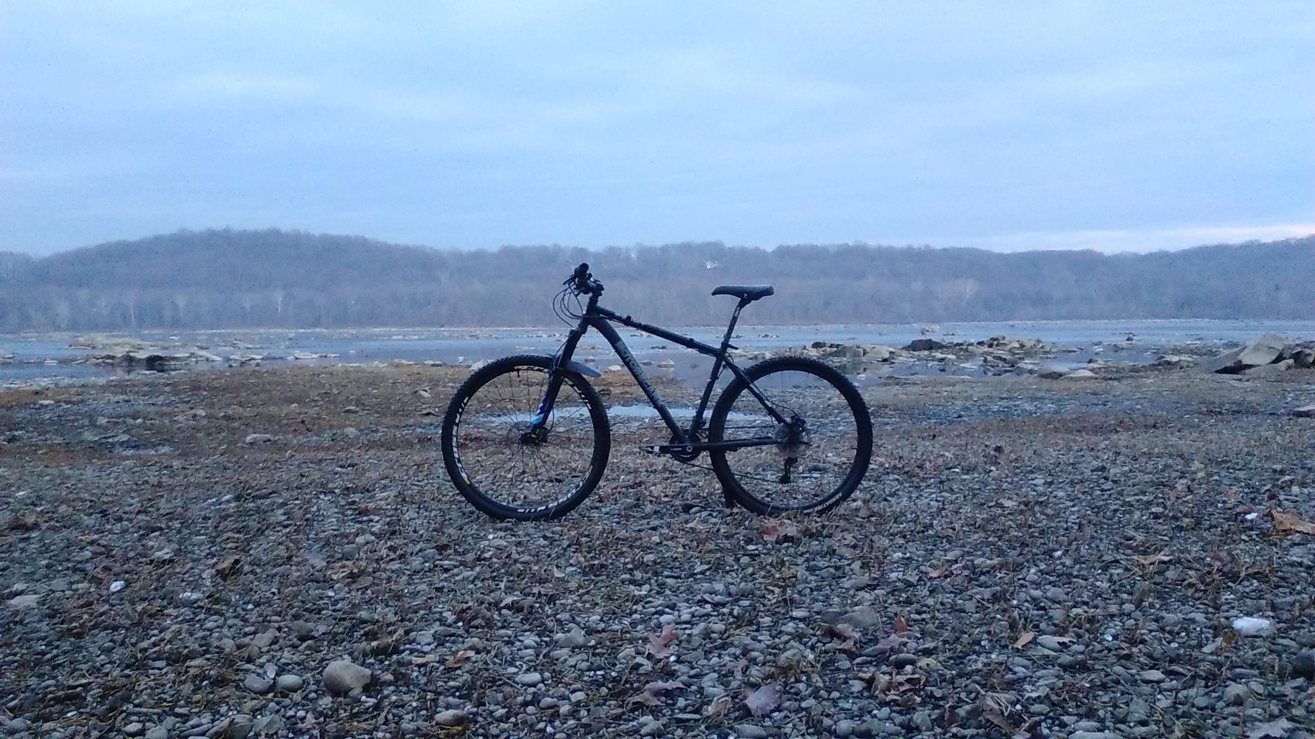 Motobecane Motobecane 529 HT: A black mountain bike parked on a rocky shoreline, with a calm river and wooded hills in the background under a cloudy sky.