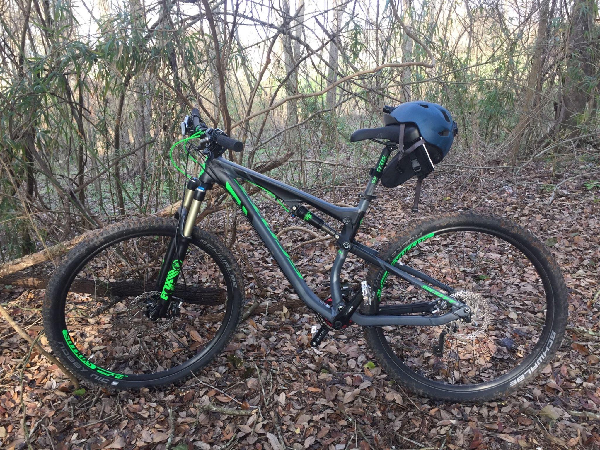 Scott Genius 920: A mountain bike with a black and green frame is parked on a bed of leaves in a wooded area. The bike has thick tires, a front suspension fork, and a black helmet attached to the seat. Surrounding the bike are trees and underbrush, indicating a natural, outdoor setting.