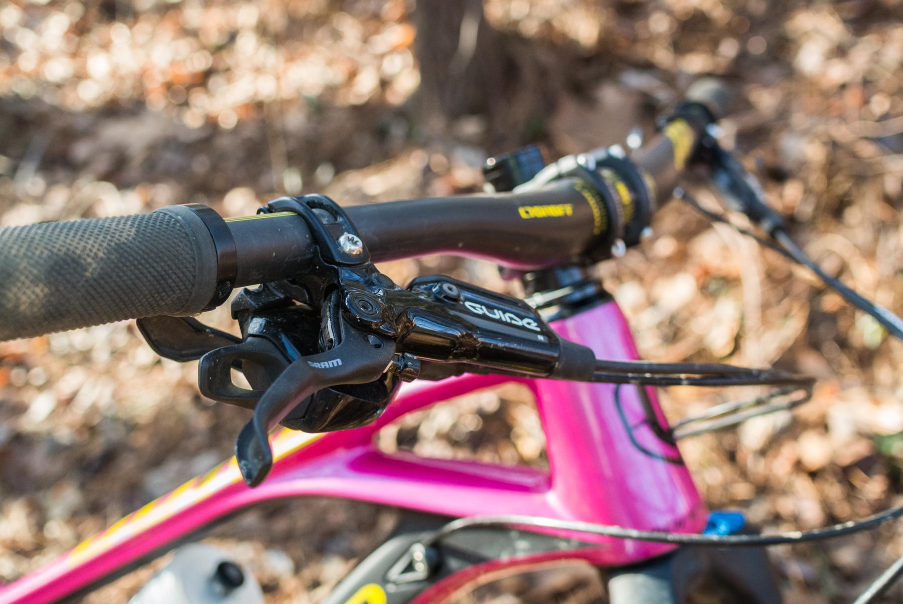 Mondraker Dune: Close-up view of a mountain bike handlebar featuring textured grips, a SRAM Guide brake lever, and colorful frame components, set against a blurred background of leaves and foliage.