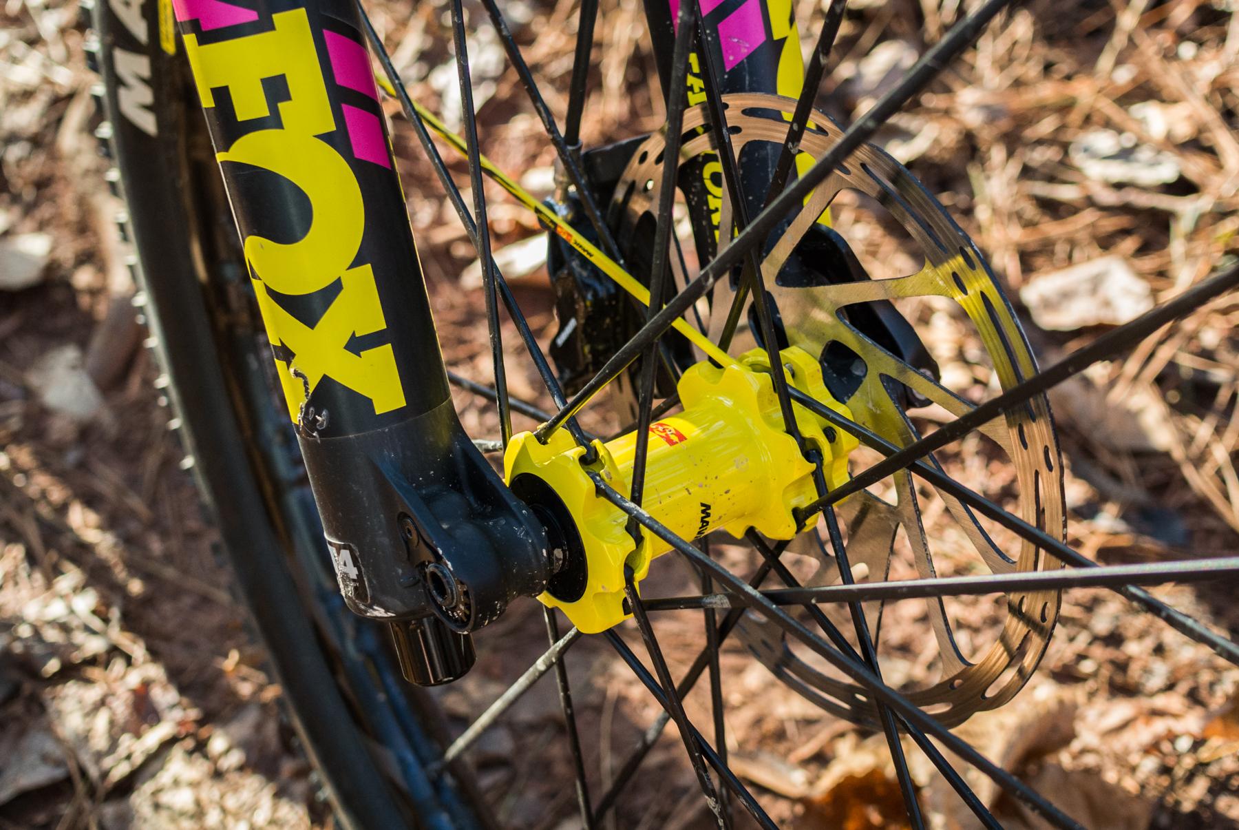Mondraker Dune: Close-up view of a bicycle wheel hub and disc brake, featuring bright yellow components and a fork marked with a large brand logo in yellow and pink. The background includes natural elements like pine needles and rocks.