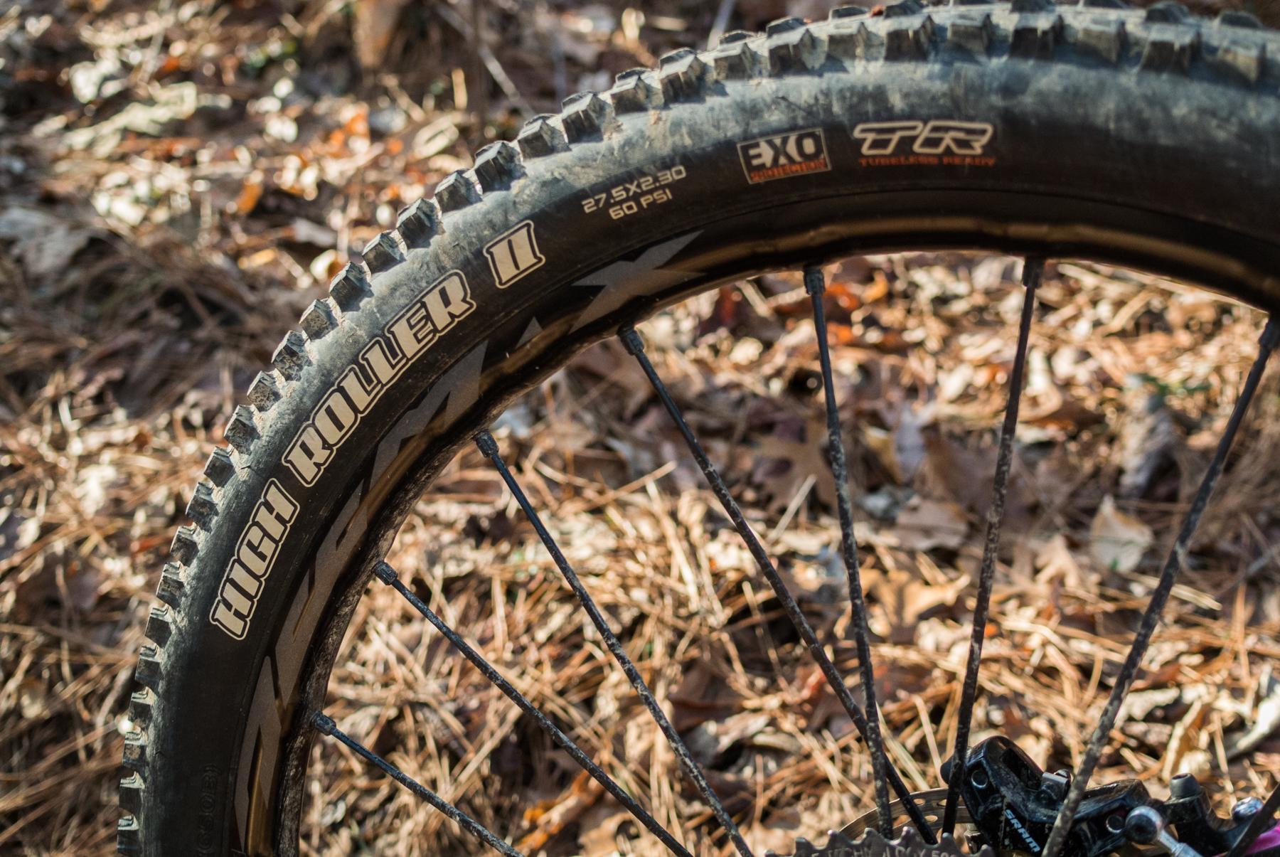Mondraker Dune: Close-up of a mountain bike tire, showcasing the tread pattern labeled "High Roller II." The tire is partially dirty and features markings indicating a size of 27.5 x 2.30 and a pressure rating of 60 PSI. The background consists of brown leaves and pine needles.