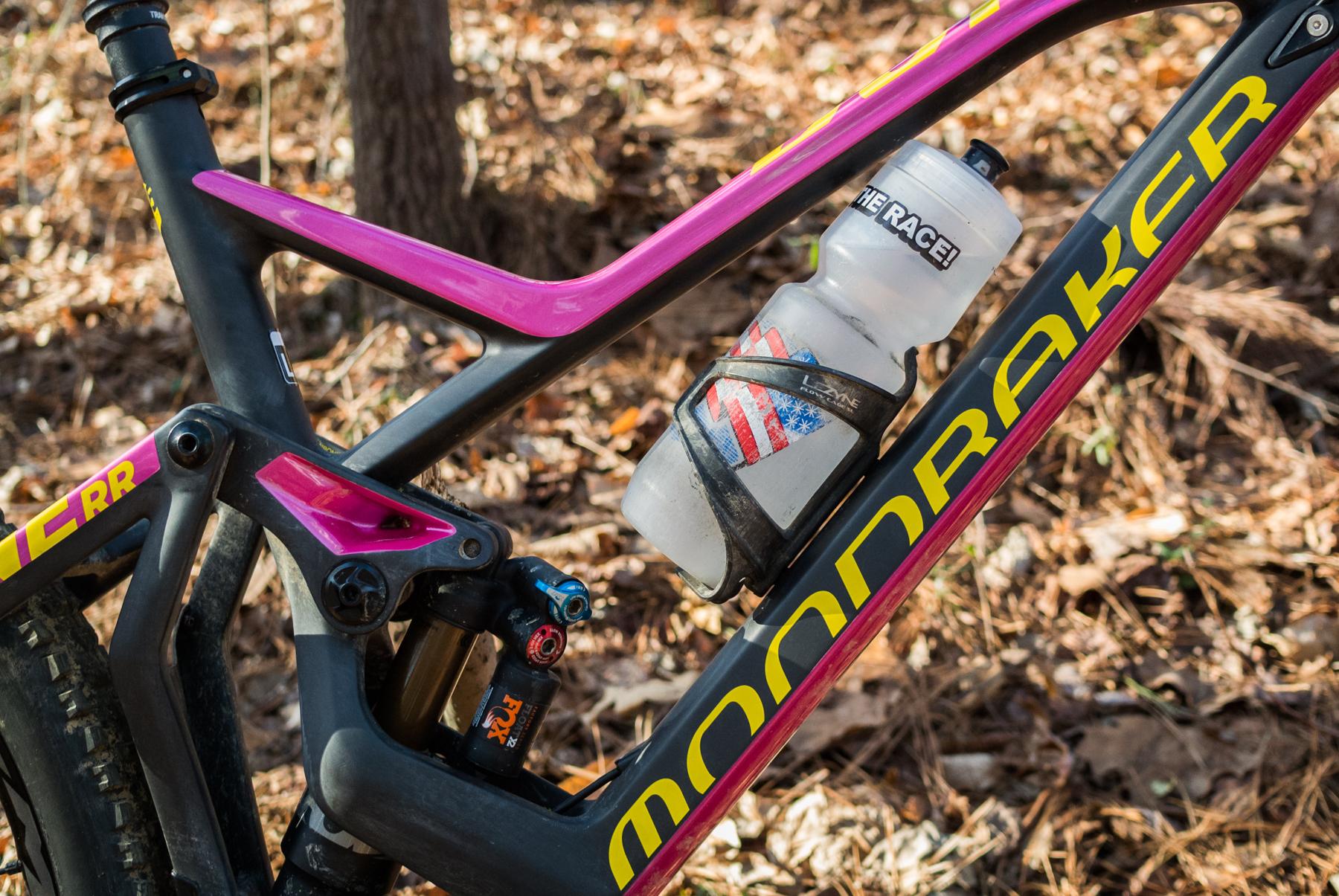 Mondraker Dune: A close-up view of a mountain bike frame featuring a black and pink design, with the word "MOONDRAKER" prominently displayed in yellow. The bike is equipped with a transparent water bottle secured in a holder, which has a design resembling the American flag. The background shows a natural setting with leaves and earth, suggesting an outdoor trail environment.