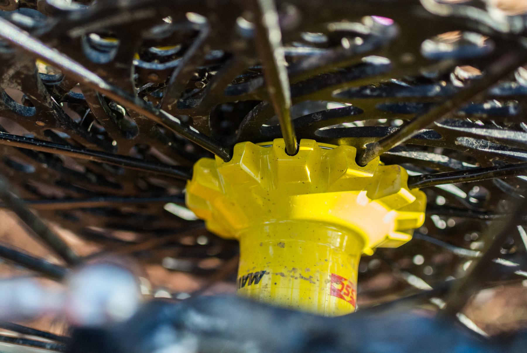 Mondraker Dune: Close-up view of a circular mechanism featuring a yellow gear at the center, surrounded by black spokes. The image highlights the intricate design of the gear system, showcasing details like ridges and connectors. The background is softly blurred, emphasizing the gear's structure.
