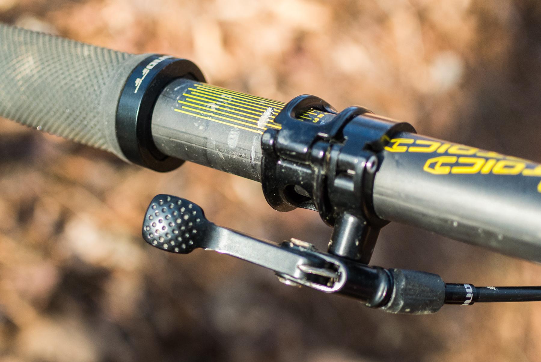 Mondraker Dune: Close-up view of a bicycle handlebar, featuring a textured grip, a locking mechanism, and a gear shifter. The handlebar is partially obscured by dirt and outdoor surroundings.