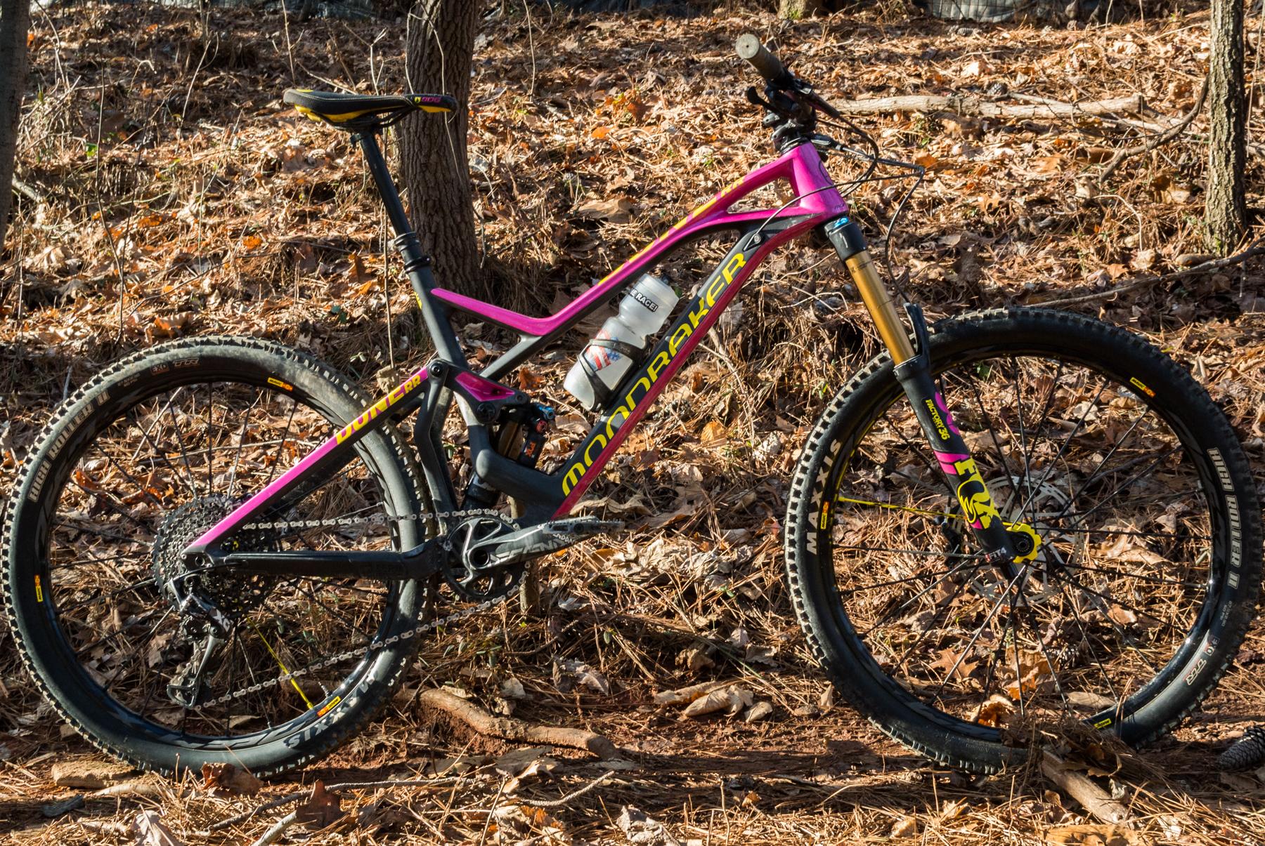 Mondraker Dune: A vibrant mountain bike with a pink and black frame, positioned on a forest floor covered in leaves and foliage. The bike features high-quality tires and a water bottle mounted on the frame, showcasing its readiness for outdoor adventures. Trees are visible in the background, indicating a natural setting.