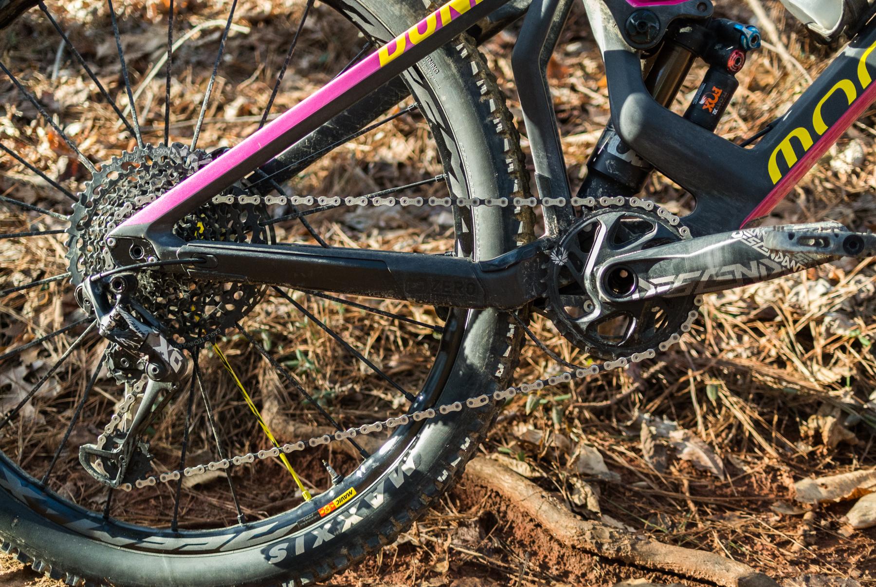Mondraker Dune: Close-up view of a mountain bike's drivetrain, featuring the rear derailleur, cassette, and chain on a brown, leaf-covered ground. The bike frame is partially visible, showcasing a black and pink design.