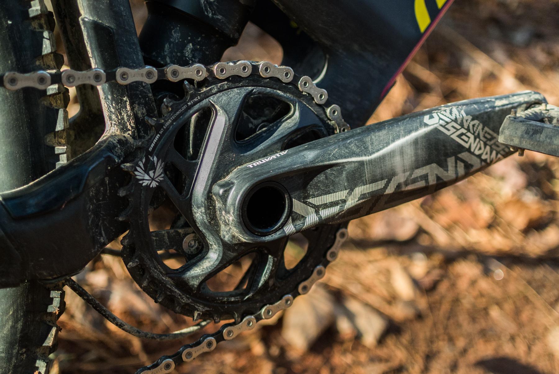 Mondraker Dune: Close-up view of a mountain bike's crankset and chain, showcasing a dirty black crank arm and chainring against a backdrop of brown dirt and rocks. The image highlights the bike's mechanical details, including the chain and the drivetrain components.