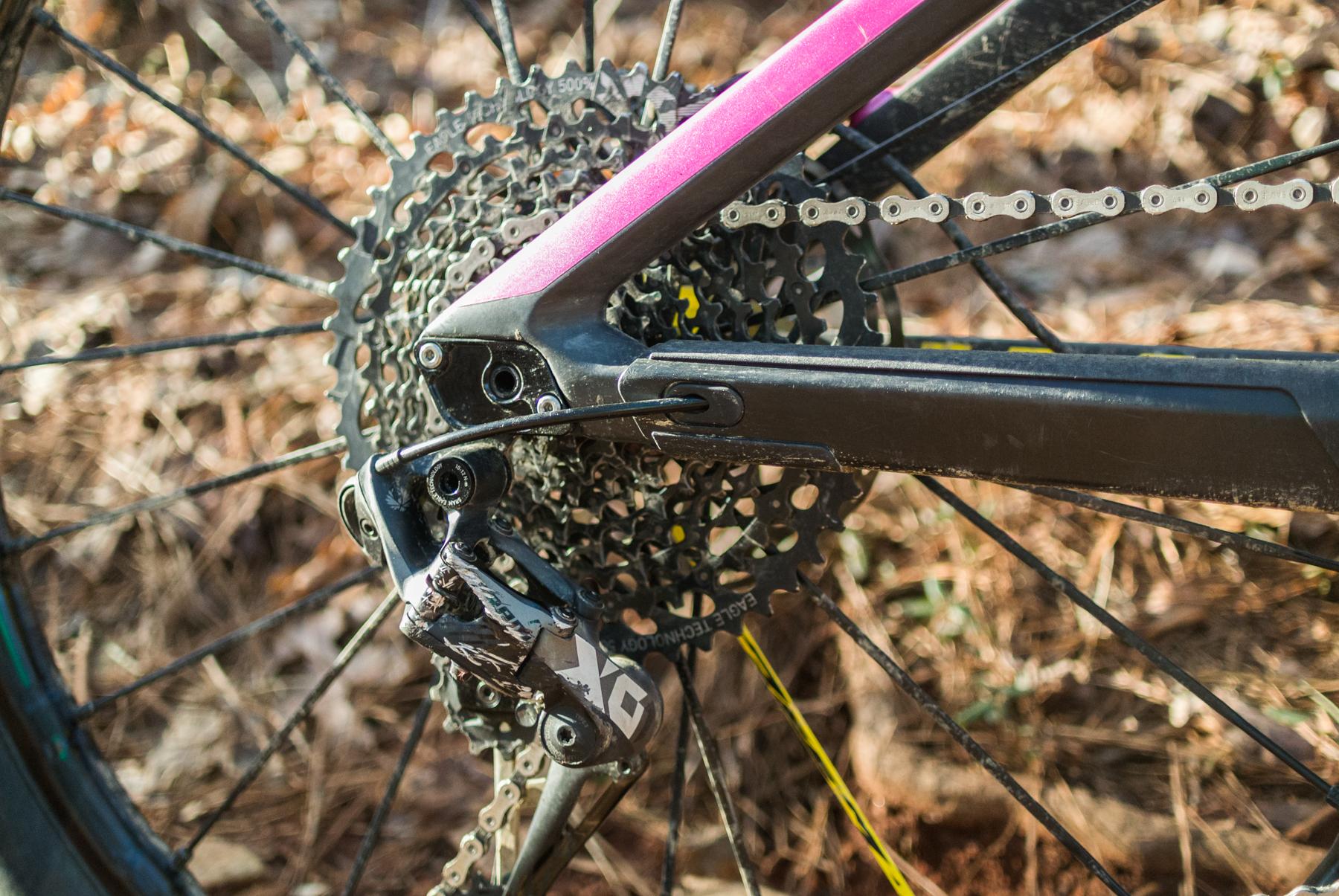 Mondraker Dune: Close-up of a bicycle's rear drivetrain, showcasing the cassette, chain, and derailleur. The image highlights a black and silver derailleur attached to a wheel with a colorful cassette, set against a natural background of leaves and dirt.