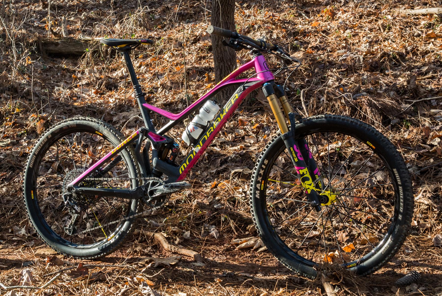 Mondraker Dune: A brightly colored mountain bike with a pink and black frame is positioned on a bed of dried leaves in a wooded area. The bike features thick tires designed for off-road terrain and has a water bottle mounted on the frame. Sunlight filters through the trees, casting soft shadows around the bike.