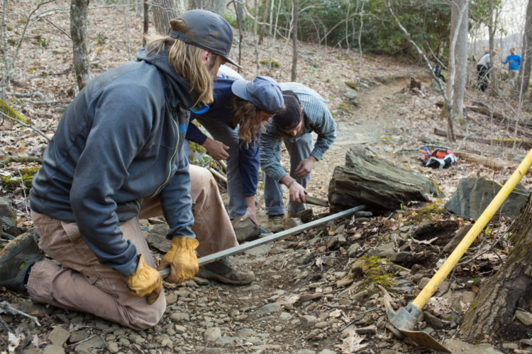 Four individuals working on a trail in a wooded area, focusing on moving a large rock. They are using tools such as a pry bar and wearing gloves while kneeling on rocky ground. In the background, additional people can be seen, along with some tools and equipment.