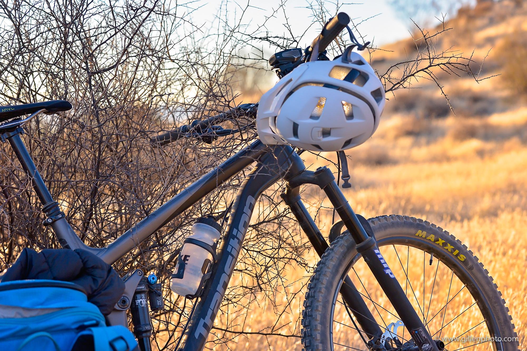 Transition Scout: A mountain bike leaned against a dry bush, with a white helmet resting on the handlebars. A blue backpack is partially visible in the foreground, and a water bottle is attached to the bike's frame. The background features golden grass and a soft, blurred landscape.