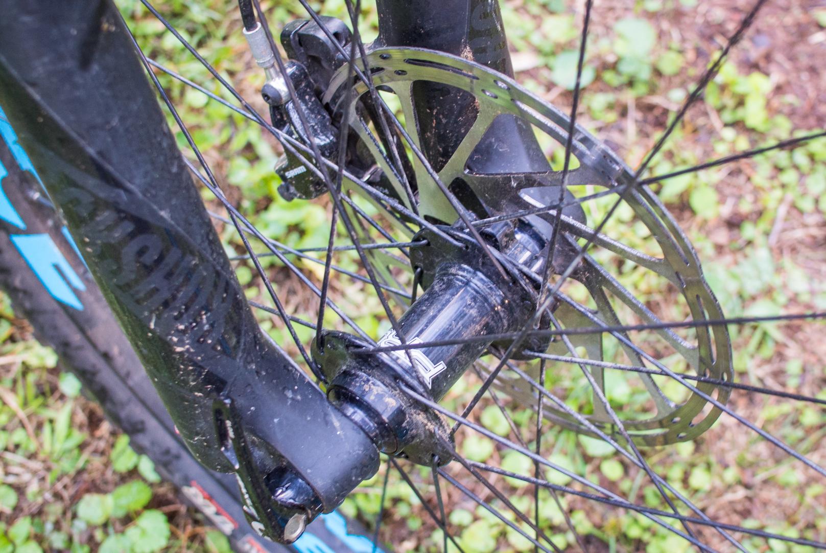 Orange Stage 5 RS: Close-up image of a bicycle front wheel hub and brake rotor, featuring visible dirt and grime, set against a background of green grass and leaves.