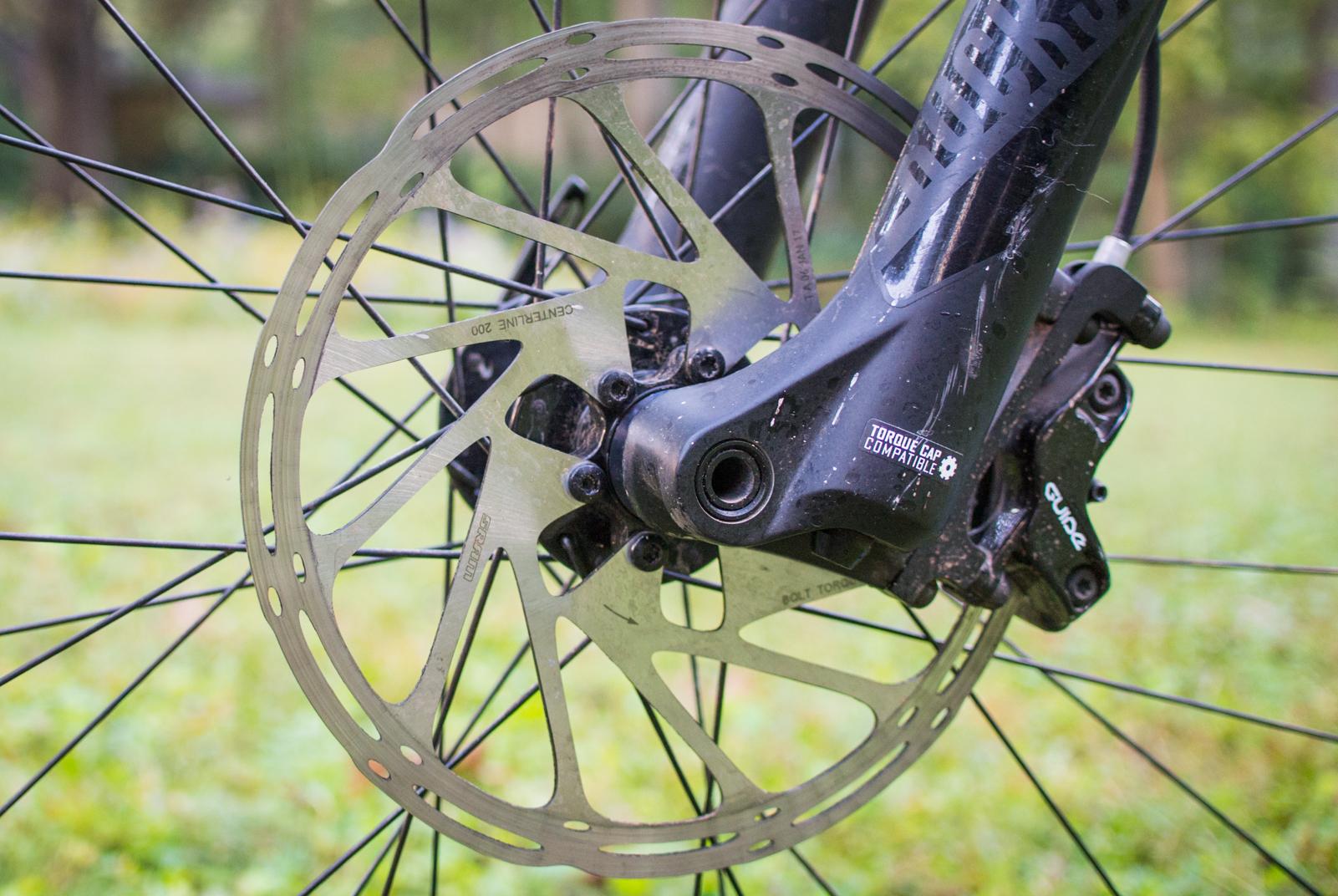 Orange Stage 5 RS: Close-up of a bicycle's front wheel featuring a disc brake system, showcasing the brake rotor and caliper attached to a black fork. The background is a blurred green natural setting.