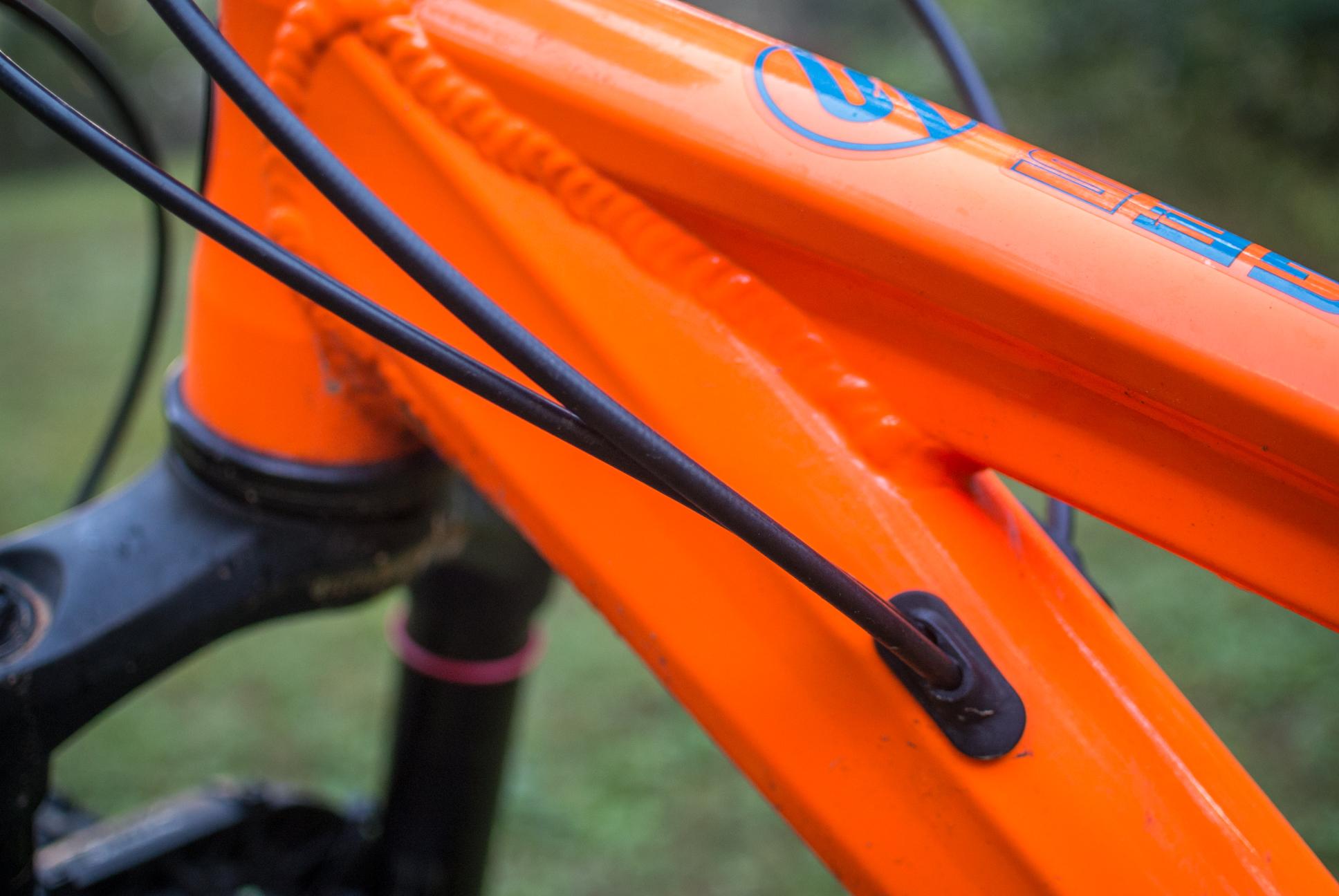 Orange Stage 5 RS: Close-up of an orange bicycle frame with black cables running along it. The image shows details of the frame's construction and a cable guide, showcasing the bike's design and maintenance features.