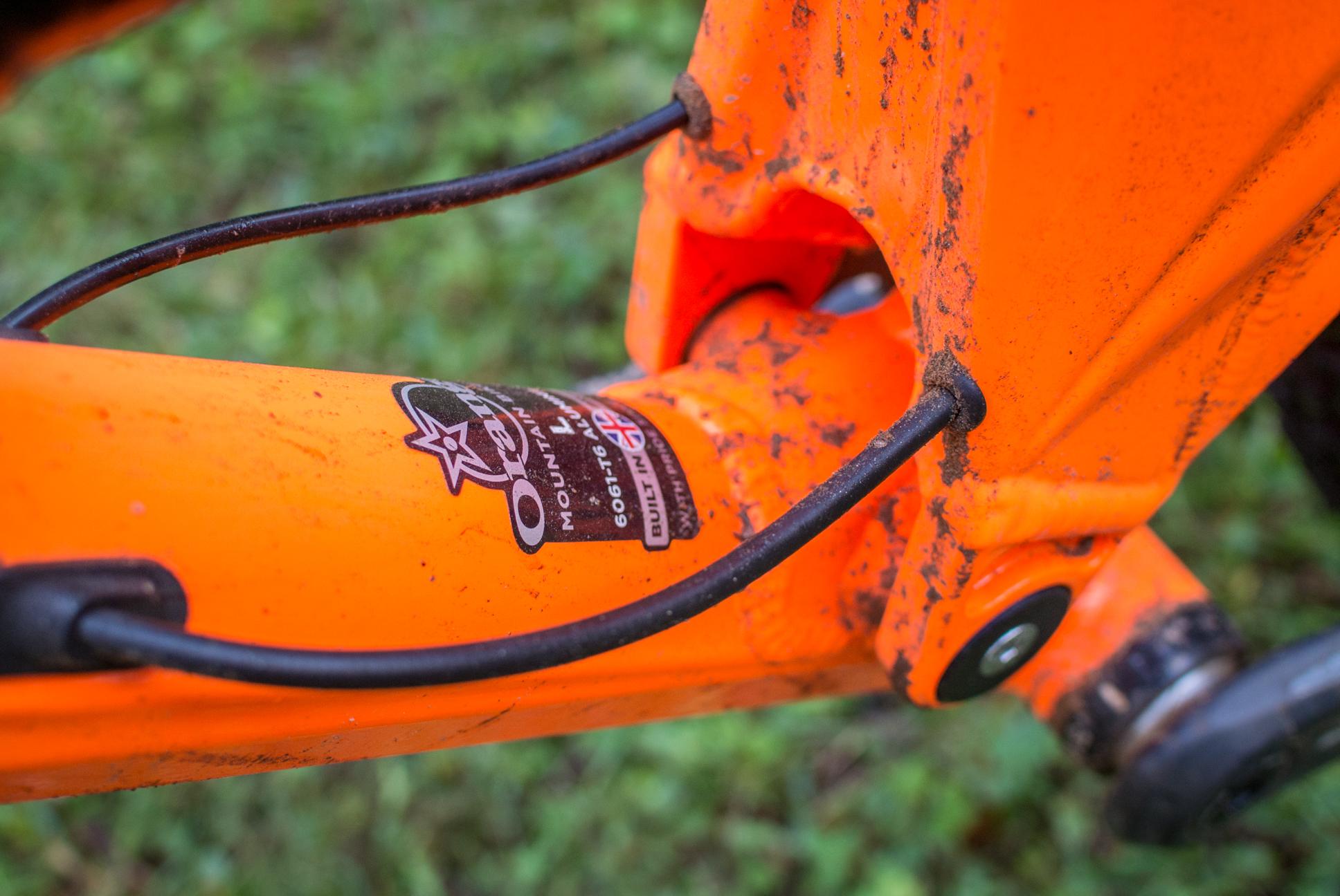 Orange Stage 5 RS: Close-up view of a bright orange mountain bike frame, showing a logo on the bottom bracket with visible dirt and grime, set against a blurred green background.