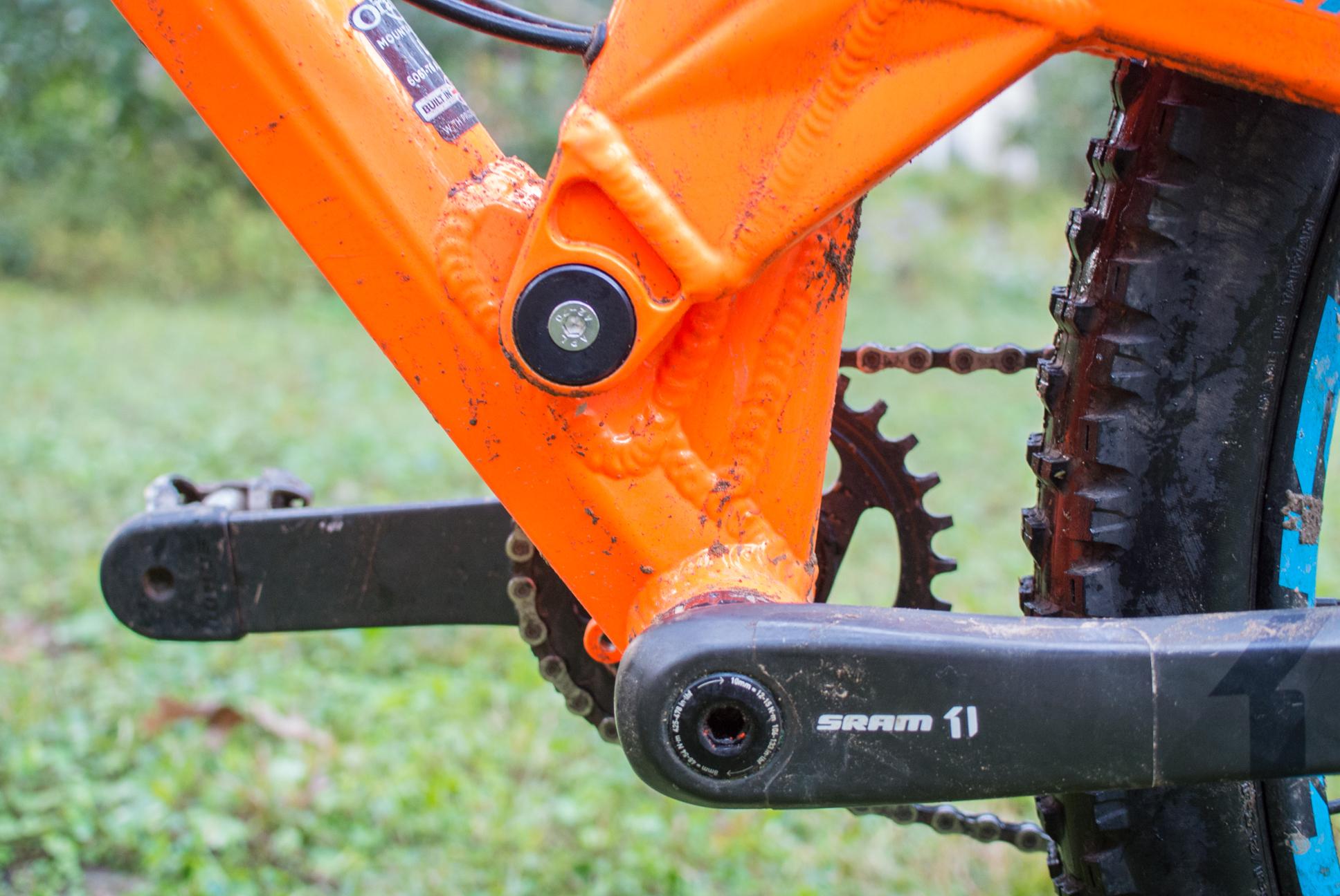 Orange Stage 5 RS: Close-up view of a mountain bike's crankset and bottom bracket area, featuring an orange frame with visible dirt and wear, a black SRAM crank, and a portion of the rear tire. The image captures the detailed components essential for bike performance and durability.