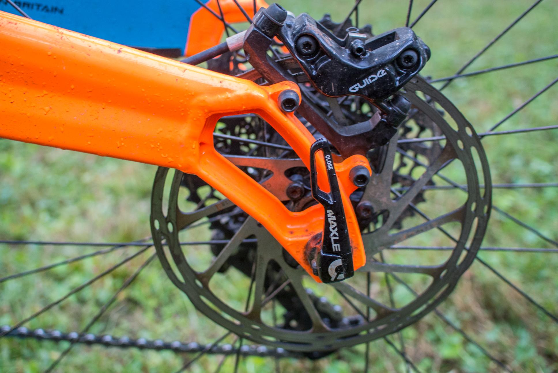Orange Stage 5 RS: Close-up view of a mountain bike's rear wheel, showcasing the orange frame, brake rotor, and derailleur components. Water droplets are visible on the frame, indicating recent exposure to rain or wet conditions. The disc brake system is detailed, including the brake caliper, alongside various mechanical parts. The background is blurred with green grass.