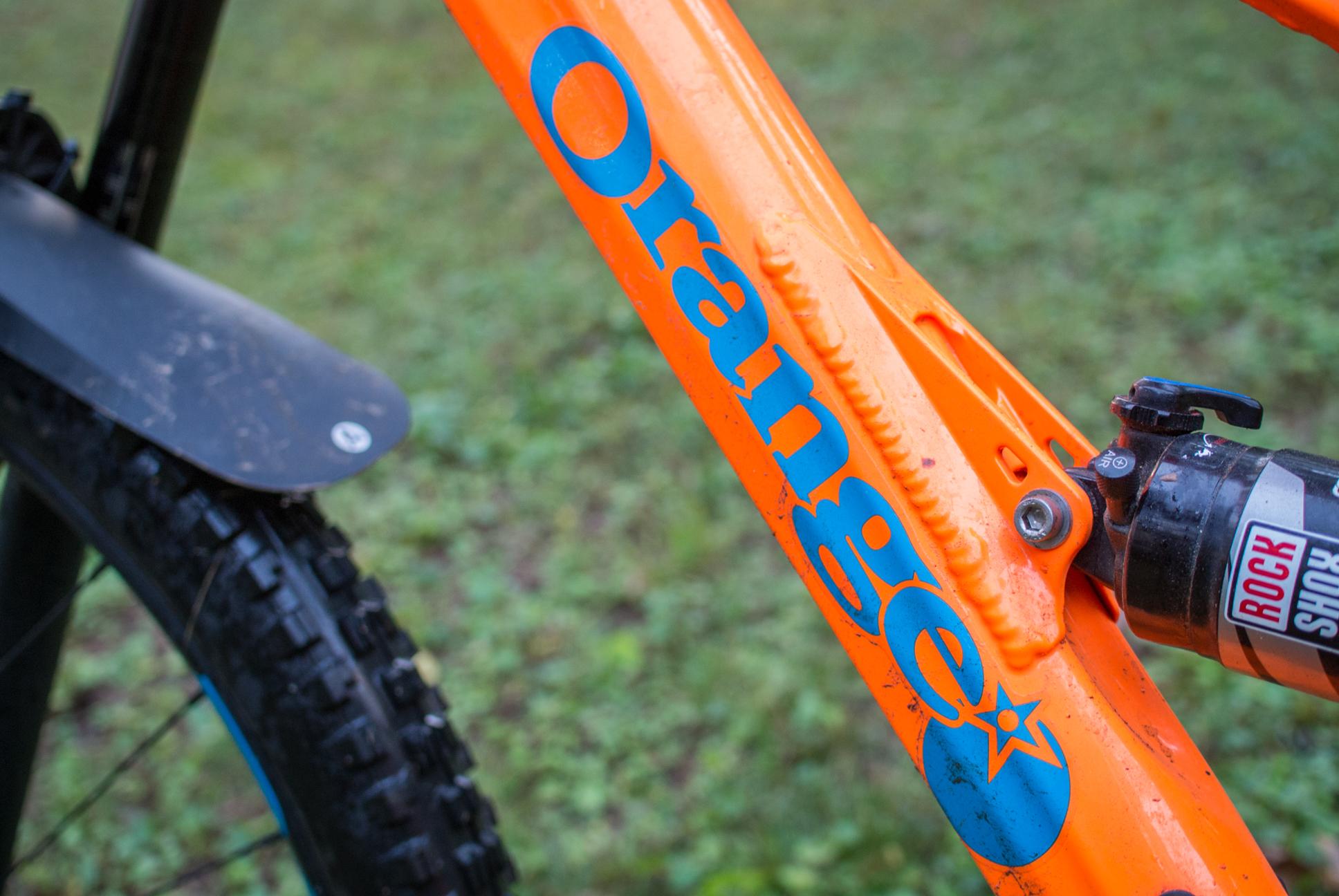 Orange Stage 5 RS: Close-up of an orange bicycle frame featuring the word "Orange" in bold blue letters, with a section of a black tire and a suspension component visible in the background. The image showcases the vibrant color and branding of the bike.