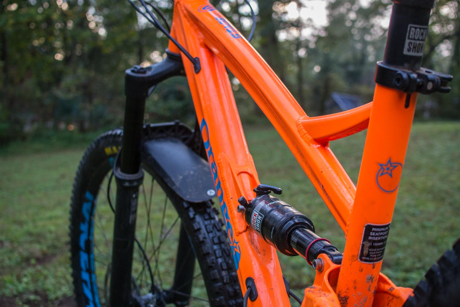 Orange Stage 5 RS: Close-up image of an orange mountain bike frame, highlighting the suspension system and components, with a blurred background of greenery. The bike features a black fork and tire with blue accents.