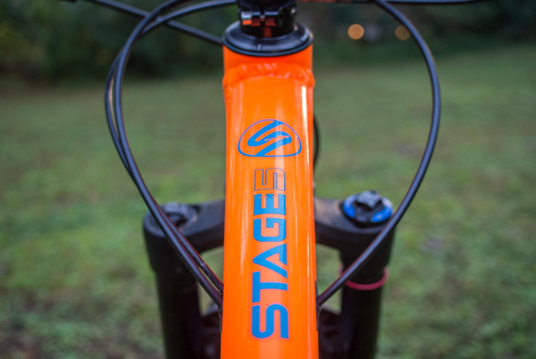 Orange Stage 5 RS: Close-up view of a vibrant orange bicycle frame featuring the "Stages" logo on the top tube, with a blurred green background. The image showcases the bike's cables and components, highlighting its design and technical features.