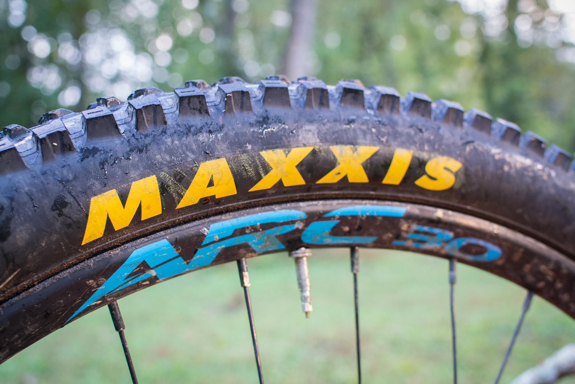 Orange Stage 5 RS: Close-up of a mountain bike tire with the brand name "Maxxis" prominently displayed in yellow and "ARC 30" in blue. The tire shows signs of use, with dirt and water droplets, and is set against a blurred green background, suggesting a natural, outdoor setting.