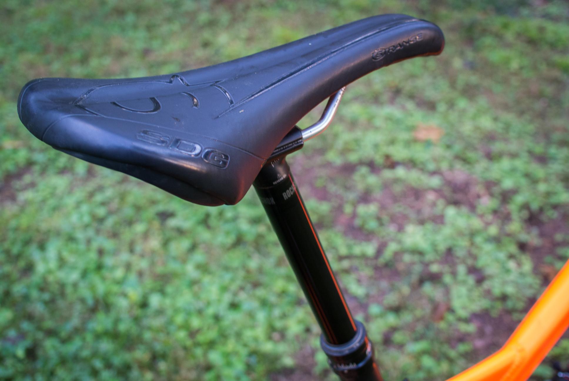 Orange Stage 5 RS: Close-up of a black bicycle saddle mounted on a seat post, with an orange bike frame partially visible in the background. The ground is covered with a patchy layer of grass.