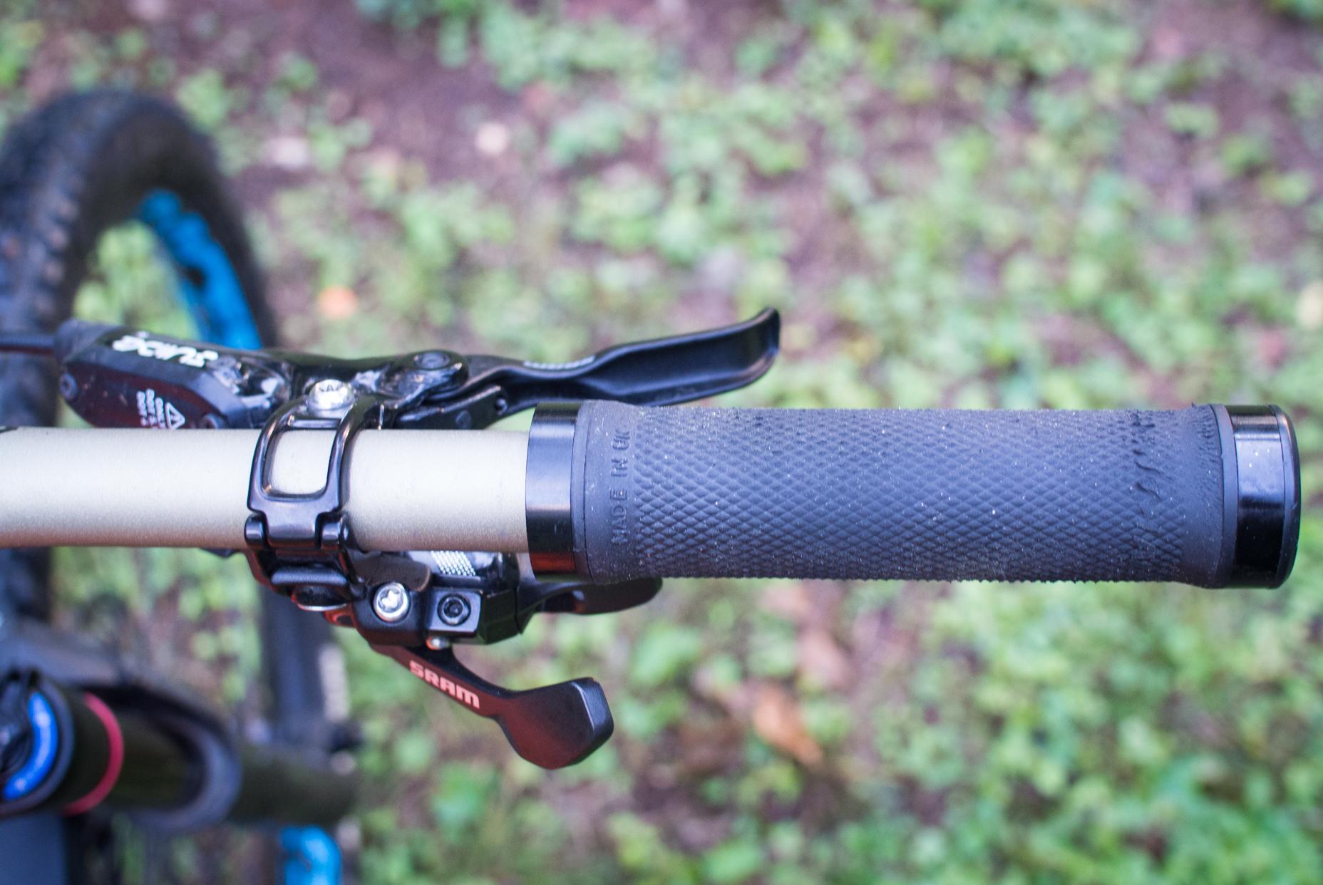 Orange Stage 5 RS: Close-up view of a mountain bike handlebar featuring a textured grip, brake lever, and gear shifter, set against a blurred natural landscape with green foliage.