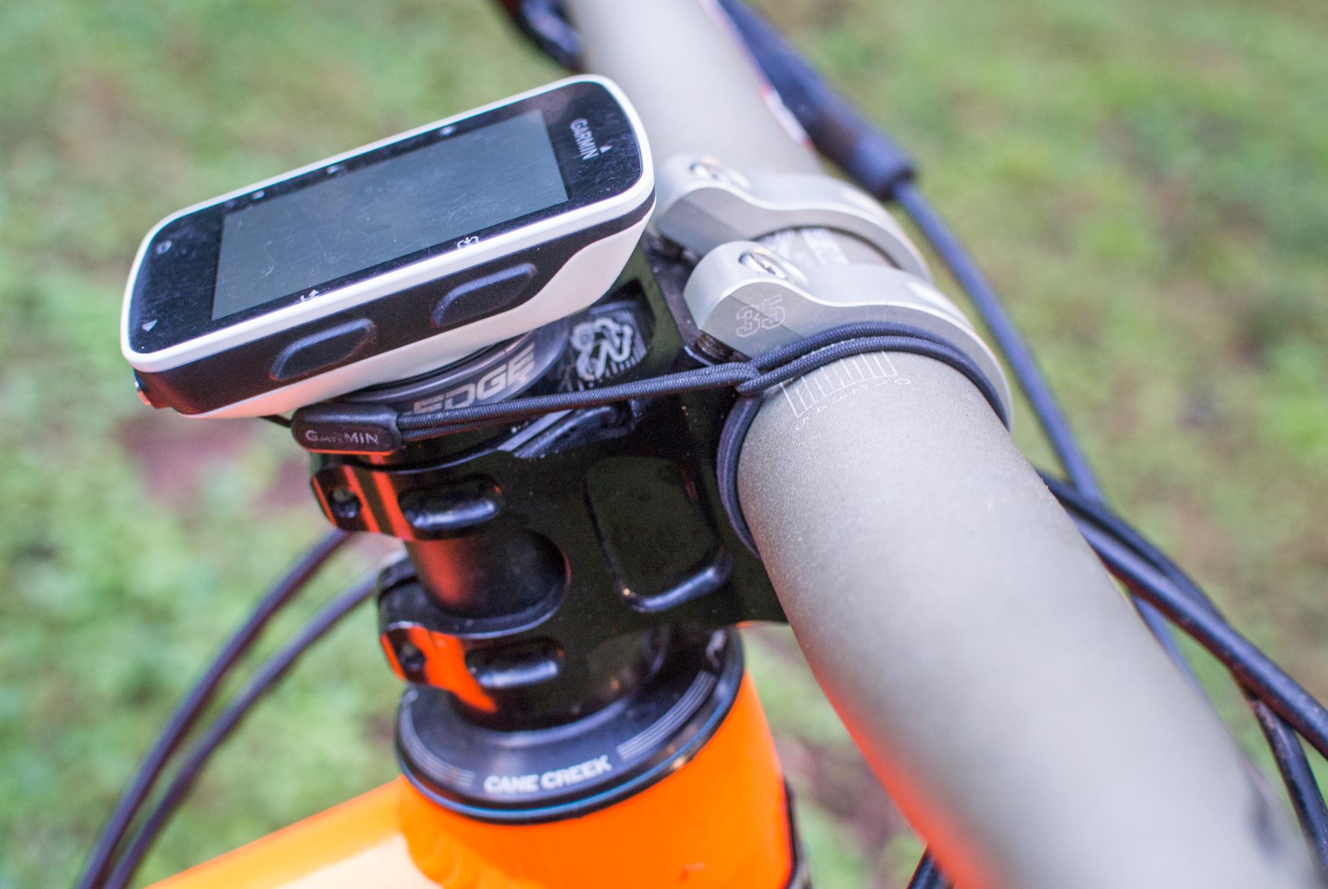 Orange Stage 5 RS: Close-up of a bicycle handlebar featuring a Garmin Edge cycling computer mounted on the stem. The handlebar is silver with a visible cable, and the bike's frame is orange in color. The background shows a blurred green surface, suggesting an outdoor setting.