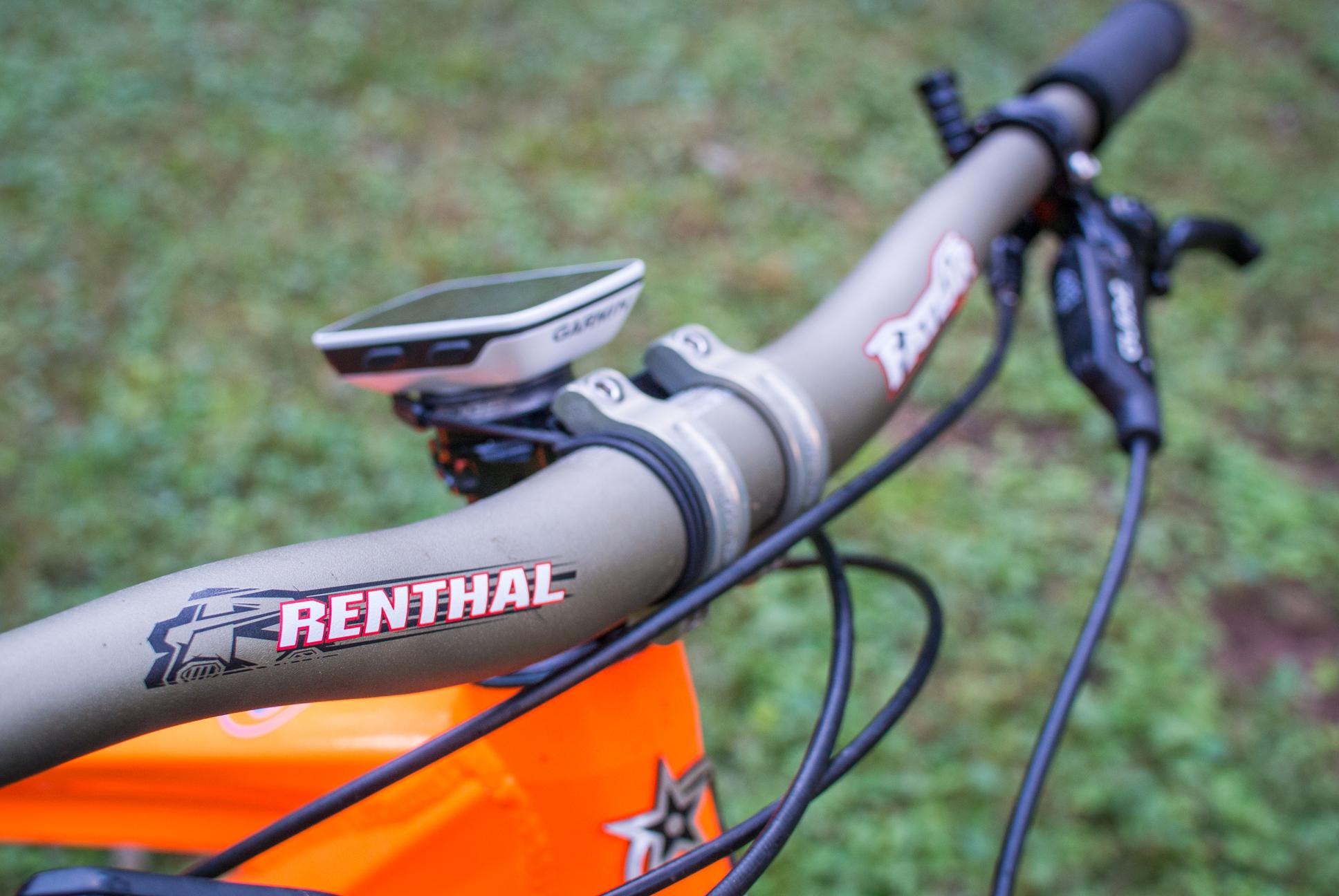 Orange Stage 5 RS: Close-up view of a bicycle handlebar featuring a Renthal logo, a cycling computer mounted on top, and a brake lever, with a blurred orange bike frame and green grass in the background.