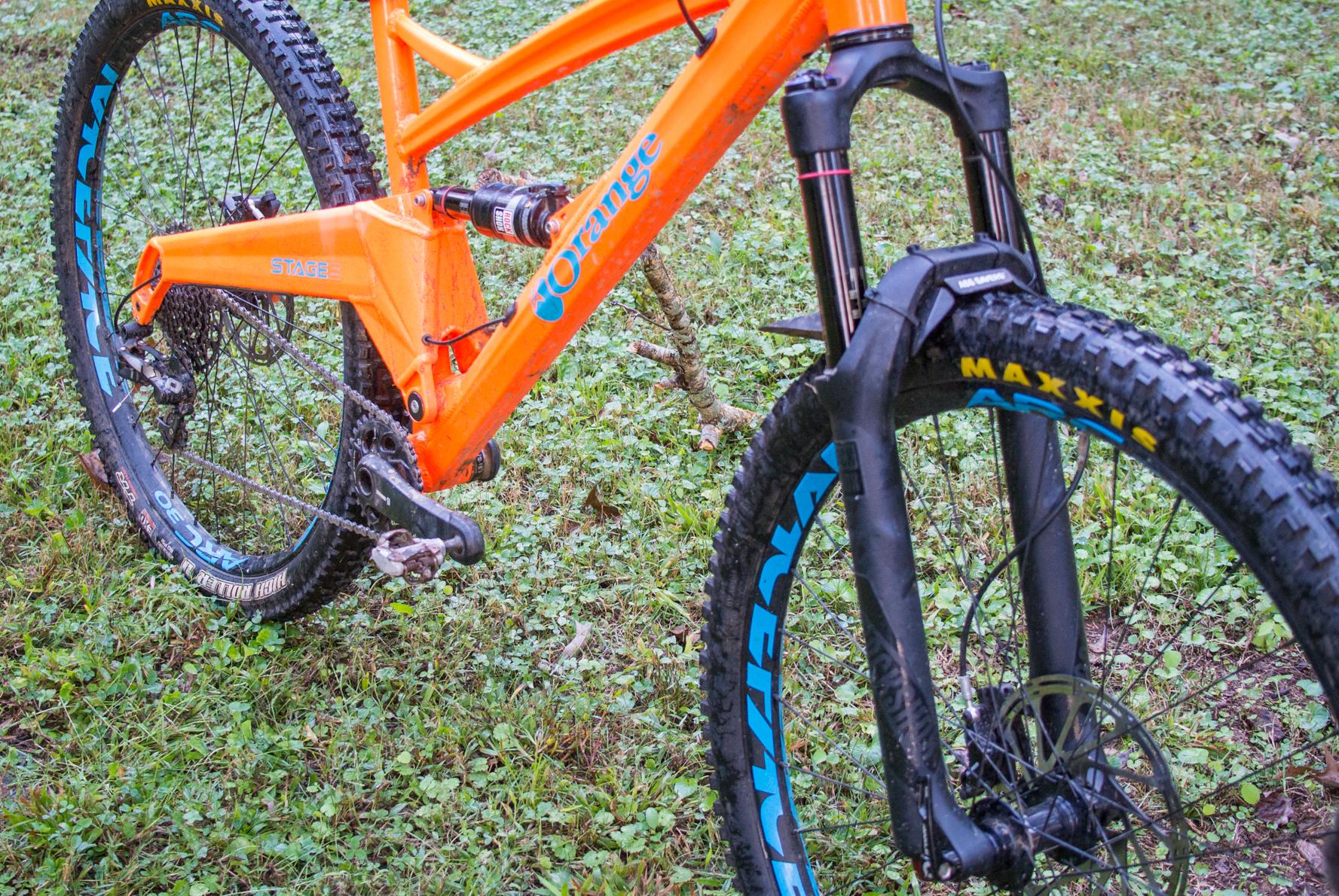 Orange Stage 5 RS: Close-up view of an orange mountain bike, showcasing the frame, suspension, and tire details on a grassy surface. The bike features prominent branding, an orange frame, black tires with blue accents, and a visible gear mechanism.
