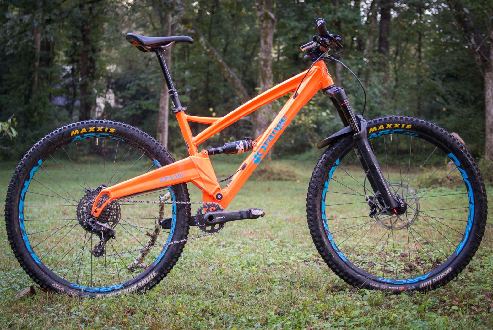 Orange Stage 5 RS: A vibrant orange mountain bike with a sturdy frame and thick tires, positioned on green grass in a wooded area. The bike features a suspension system, wide handlebars, and disc brakes, with "Maxxis" branding visible on the tires. The background includes blurred trees, indicating a natural setting.
