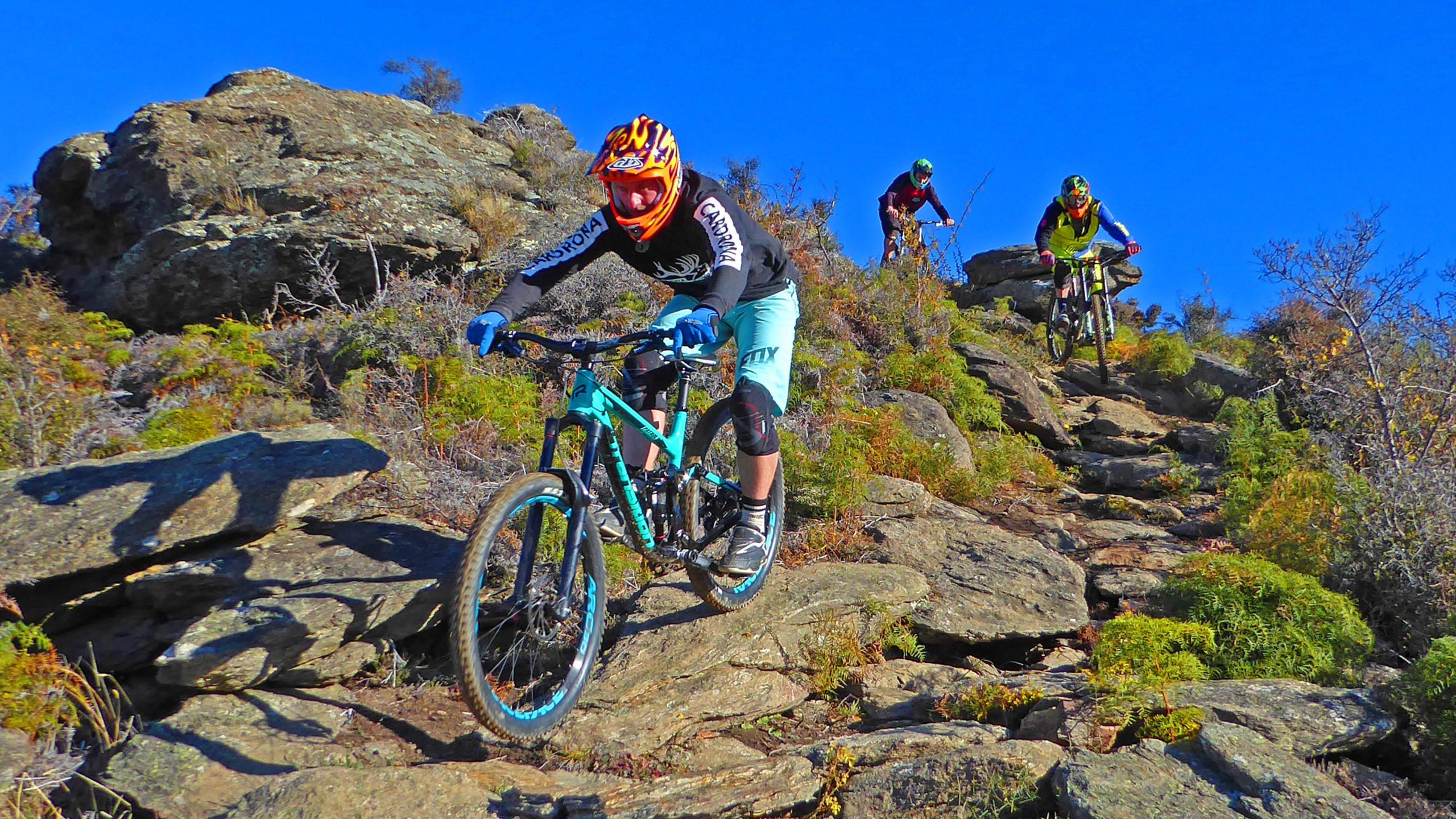 Three mountain bikers navigating a rocky trail surrounded by sparse vegetation against a clear blue sky. The front rider is in a bright helmet and gear, focused on descending the rugged path, while the other two riders are further back, also maneuvering their bikes over the stones. Linger and Die mountain bike trail.