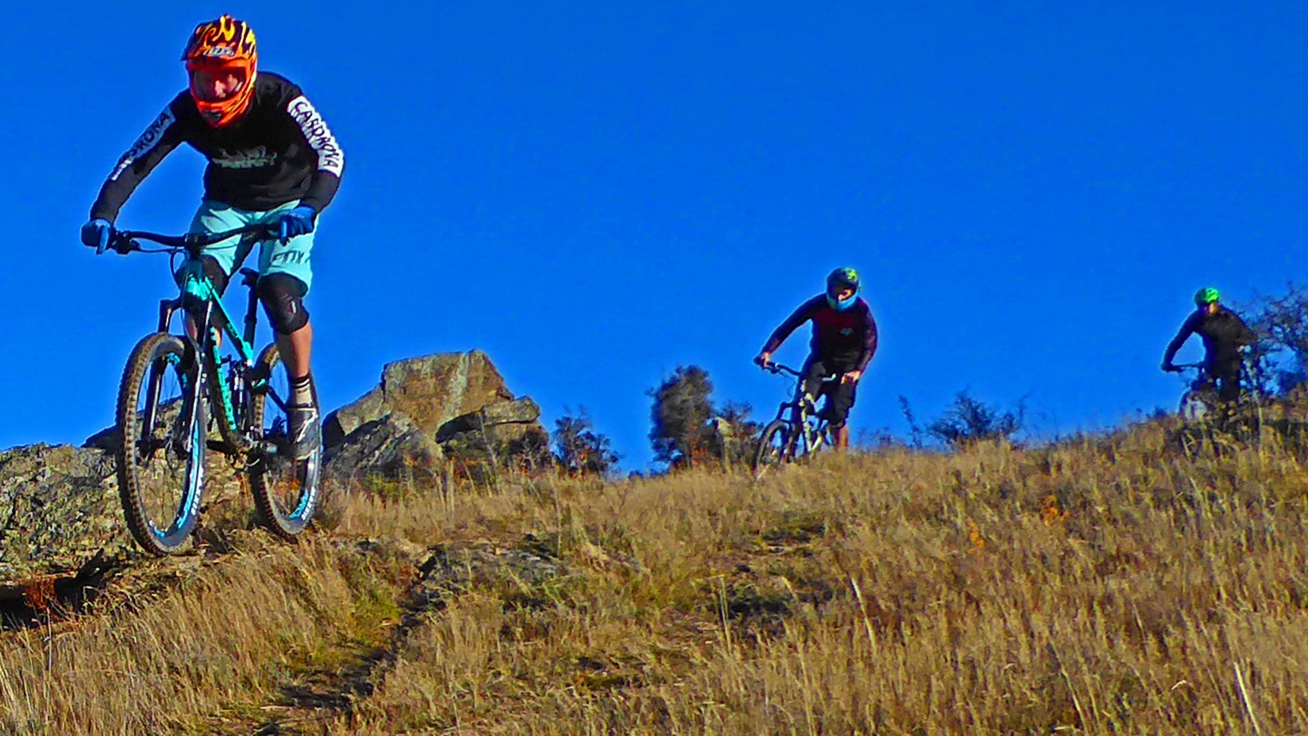 Three mountain bikers navigate a hilly, rocky trail under a clear blue sky. The lead rider, wearing an orange helmet and a black long-sleeve shirt, is seen in mid-action on a turquoise bike. The other two riders follow closely behind, dressed in darker attire with bright green helmets. The ground is covered in dry grass and rocky outcrops, creating a rugged outdoor setting. Linger and Die mountain bike trail.