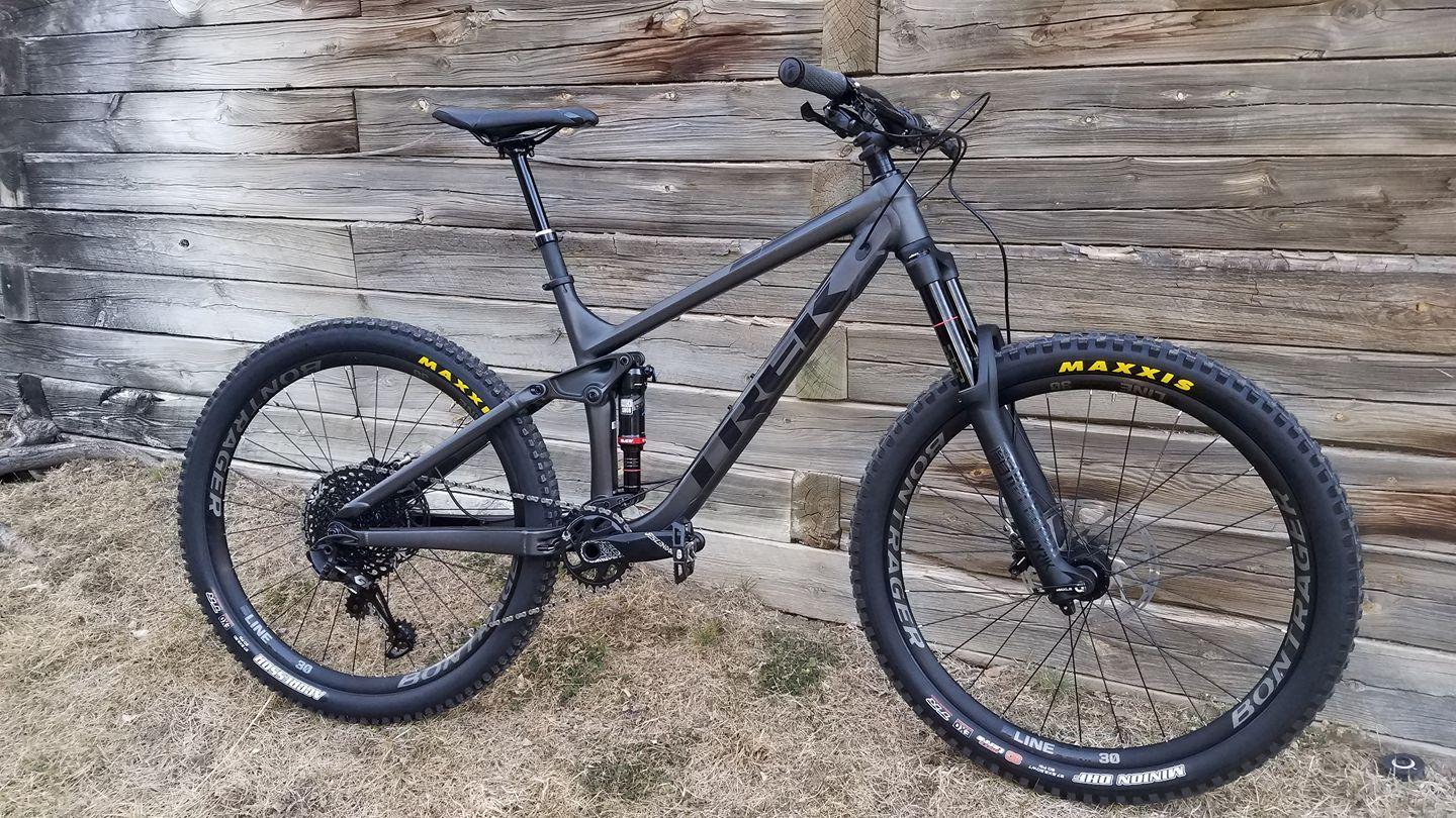 Trek Trek Remedy 8 27.5: A black mountain bike leaning against a rustic wooden wall, featuring prominent tires and high-quality components. The bike is designed for off-road use, with suspension visible and branded wheels, showcasing its rugged construction and ready-for-adventure aesthetic.