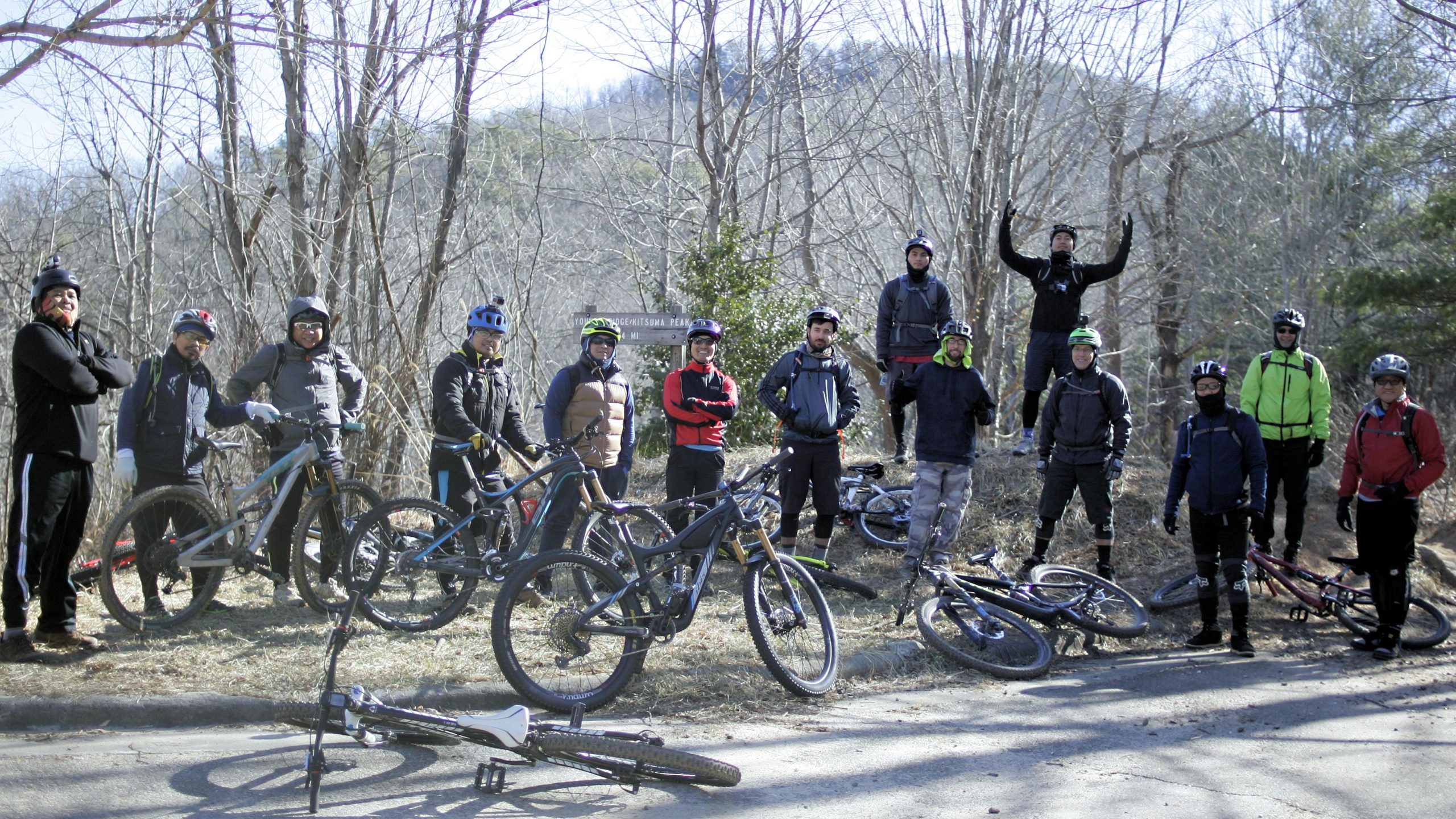 A group of mountain bikers posing together outdoors, surrounded by bare trees and a hilly background. Some bikers are standing with their bikes, while a couple are seated on the ground. The scene captures a sense of camaraderie and enthusiasm for biking. Kitsuma mountain bike trail.