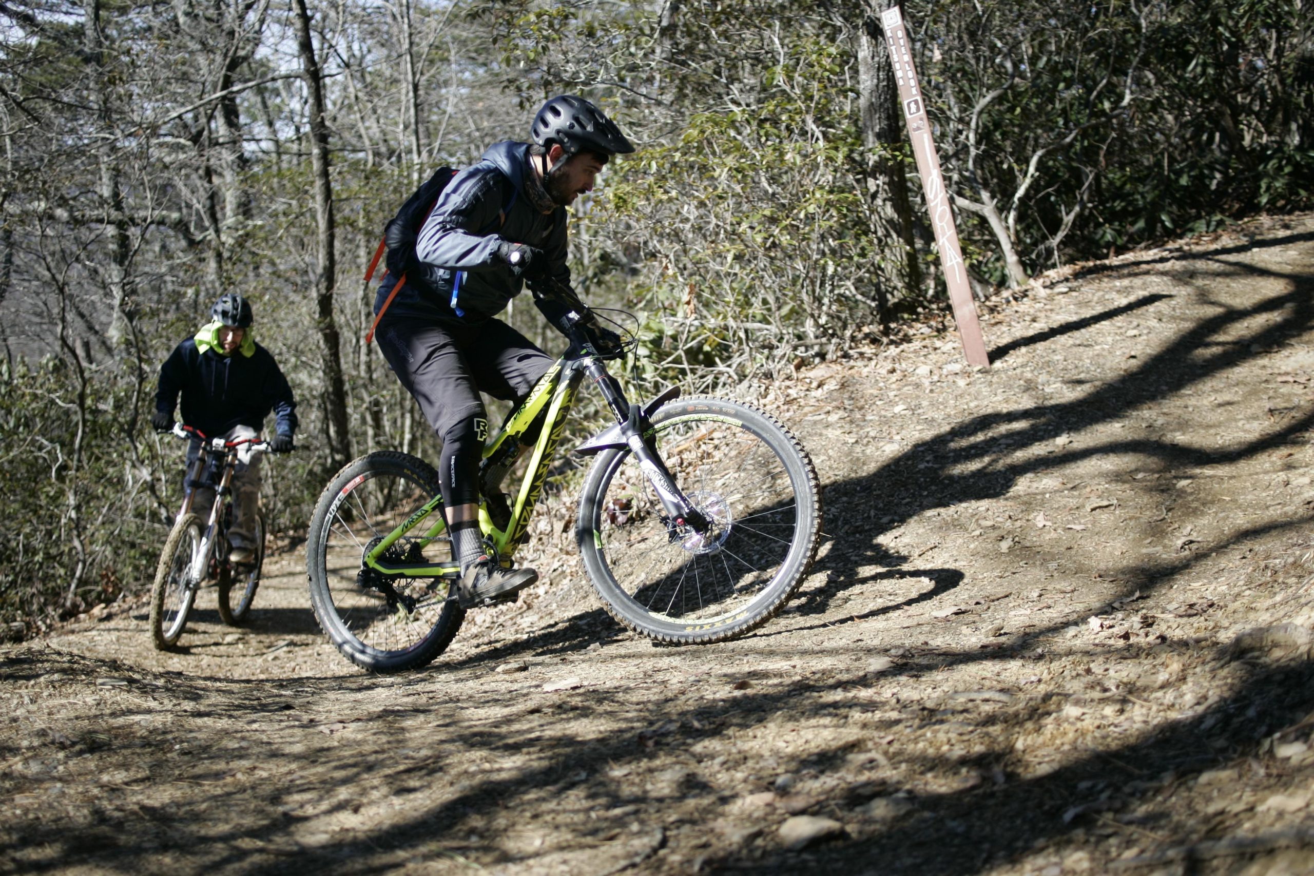 Two mountain bikers navigating a forested trail in a hilly area. One rider is approaching from the background, while the other is in the foreground, leaning forward as they navigate a steep incline. Trees and shrubs line the path, which has a mix of dirt and small rocks. A trail marker is visible in the background. Kitsuma mountain bike trail.