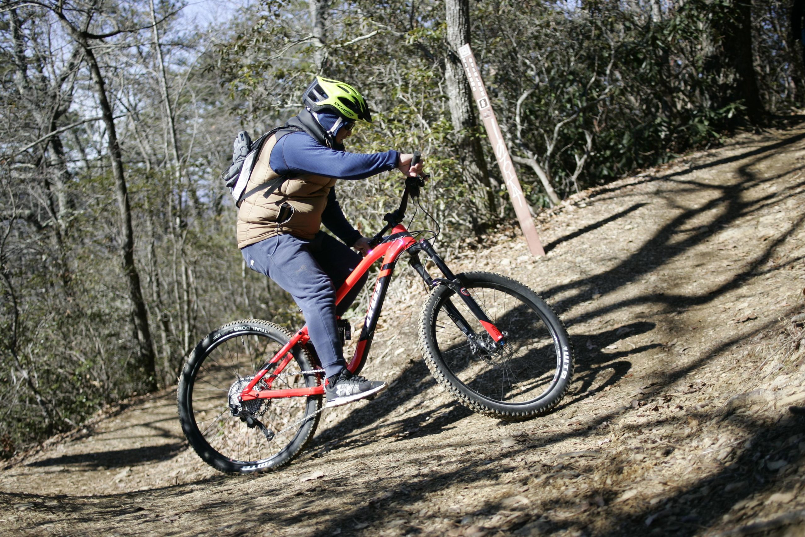A person riding a red mountain bike along a dirt trail in a wooded area. The rider is wearing a green helmet and has a backpack. A trail sign is visible in the background, indicating the direction of the path. Sunlight filters through the trees, creating a vibrant outdoor scene. Kitsuma mountain bike trail.