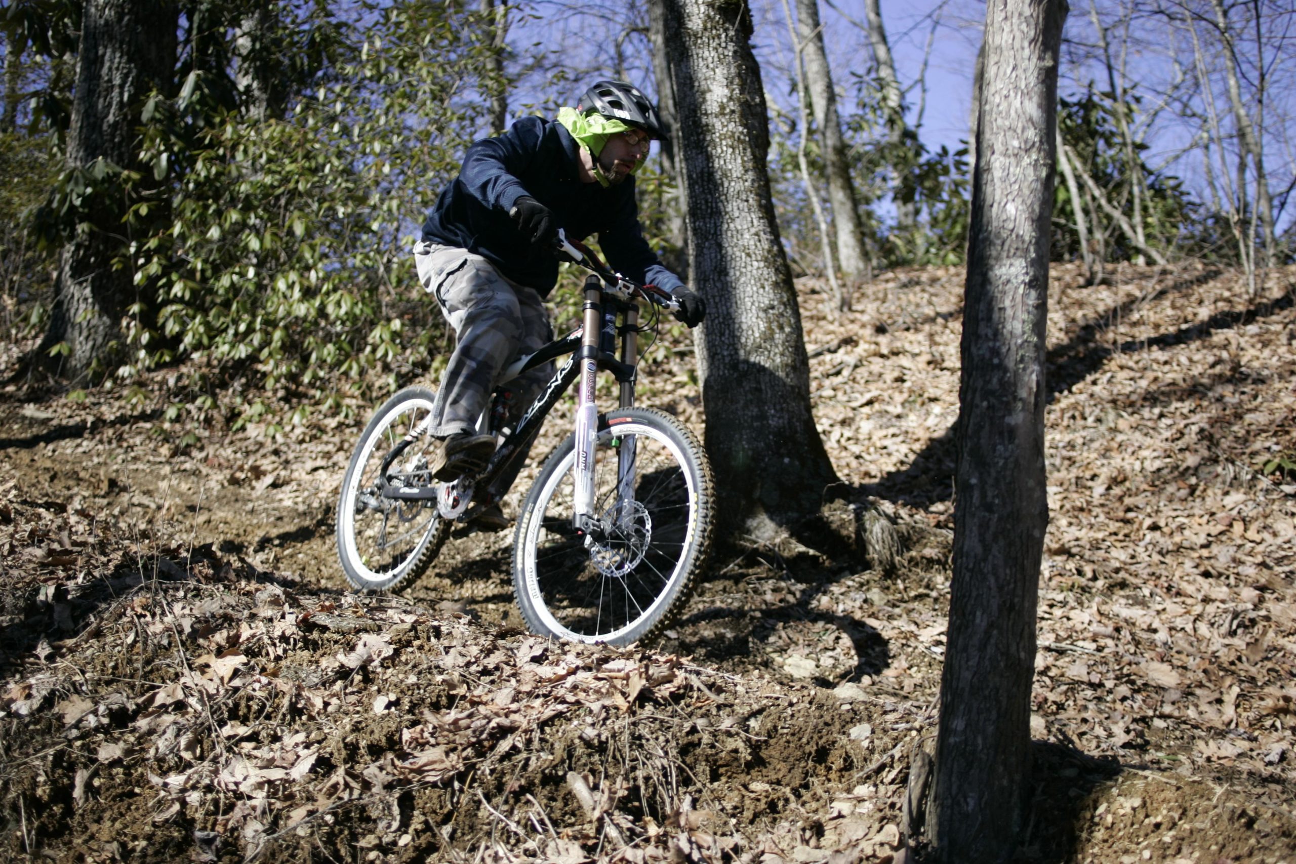 A mountain biker navigating a rocky trail in a wooded area, surrounded by trees and fallen leaves. The cyclist is wearing a helmet and a jacket, showcasing an action shot as they lean forward on their bike. Kitsuma mountain bike trail.