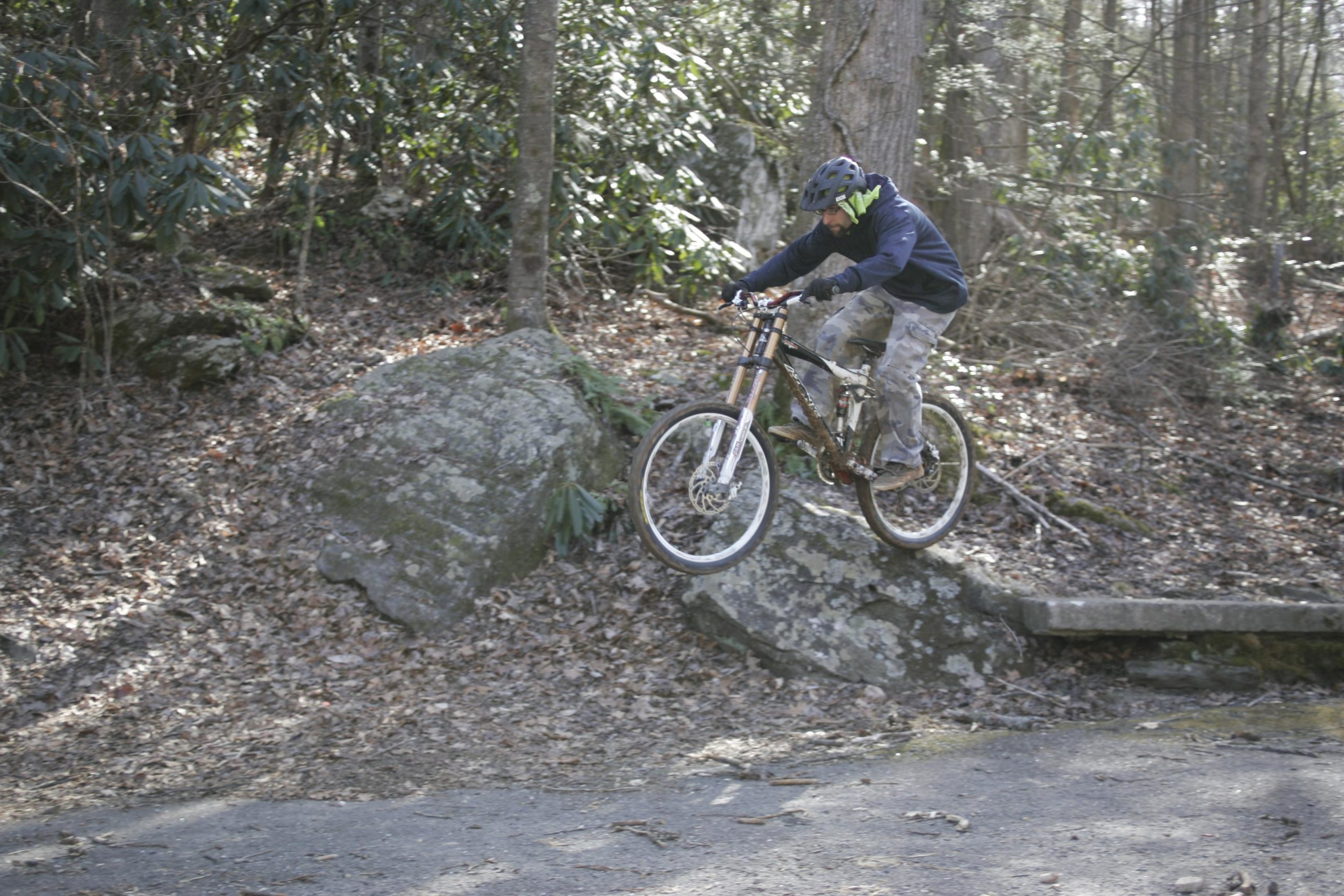 A mountain biker wearing a helmet and a dark shirt jumps off a rock onto a dirt trail in a wooded area, surrounded by trees and fallen leaves. Kitsuma mountain bike trail.