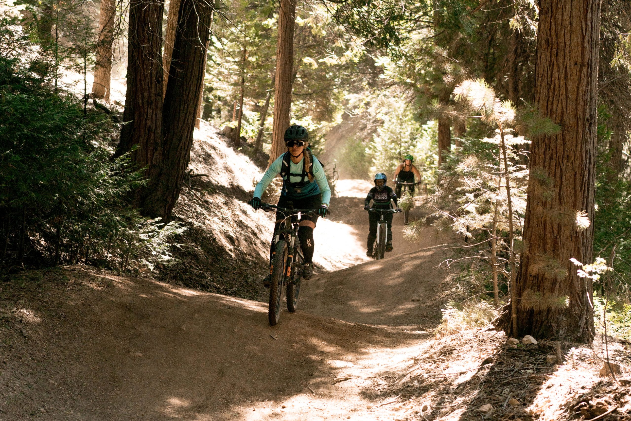 A group of three mountain bikers riding through a forested trail. The first rider, wearing a blue long-sleeve shirt and helmet, is in the foreground, while two other cyclists are visible in the background, navigating the dusty path surrounded by tall trees and sunlight filtering through the foliage. Sleigh Ride mountain bike trail.