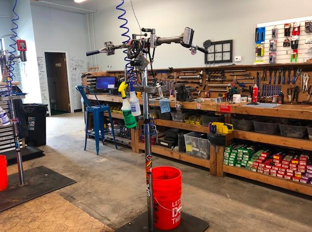 A workshop interior featuring various tools and equipment. In the foreground, there are two bike repair stands with tools attached. A bright red bucket is positioned nearby. The backdrop includes a wooden workbench laden with an assortment of tools, cleaning supplies, and a computer, against a wall lined with additional tools. The floor is concrete, and the overall atmosphere is organized and functional.